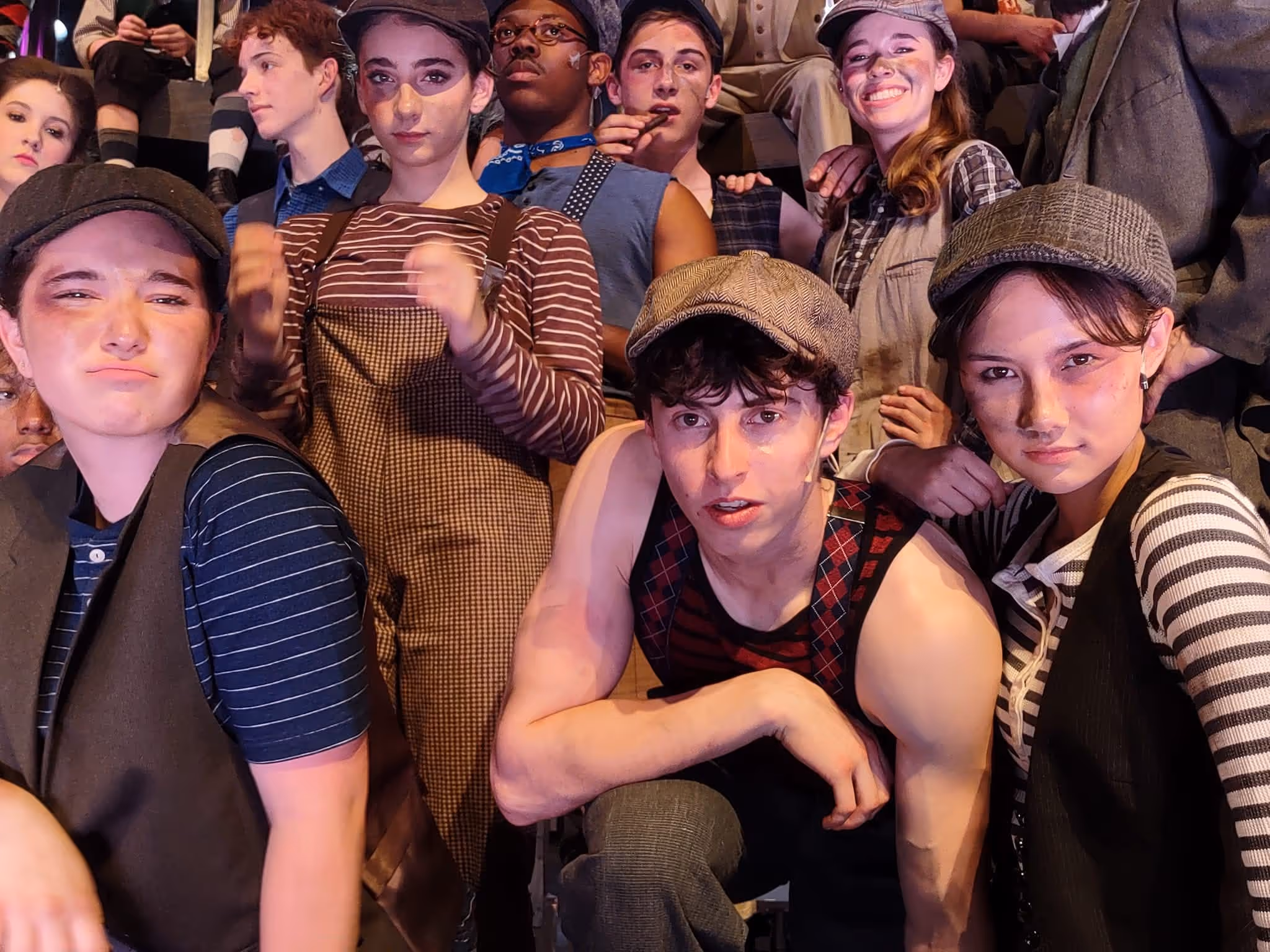 Group of young people dressed in vintage newsboy attire, posing closely together with expressive faces under stage lighting.