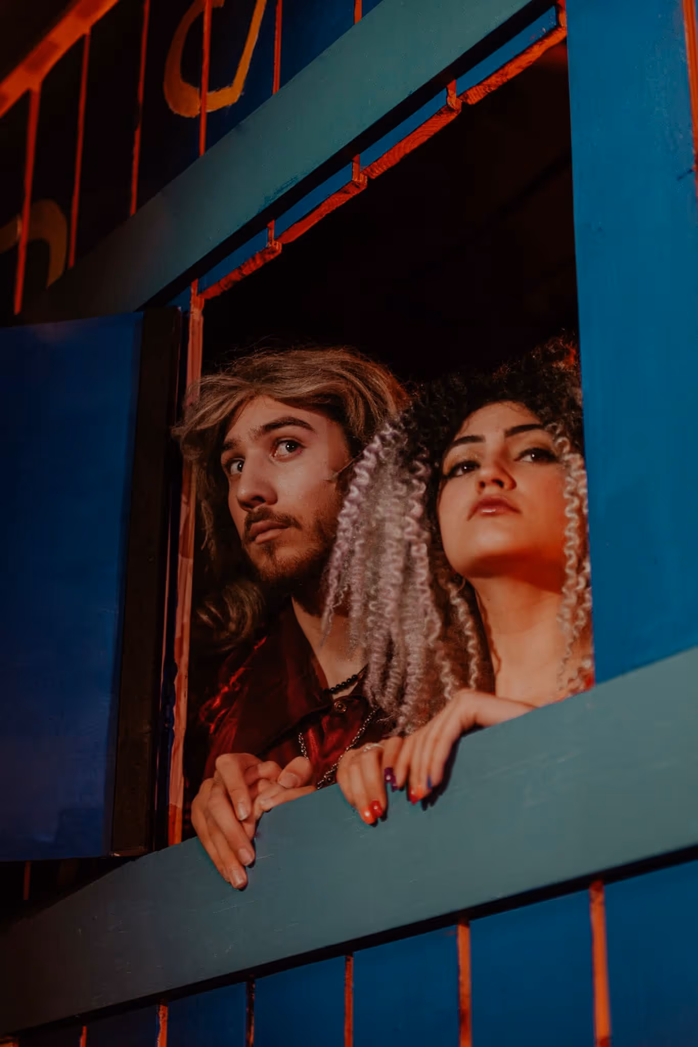 A man with long hair and a beard and a woman with curly hair leaning out of a blue window frame at night.