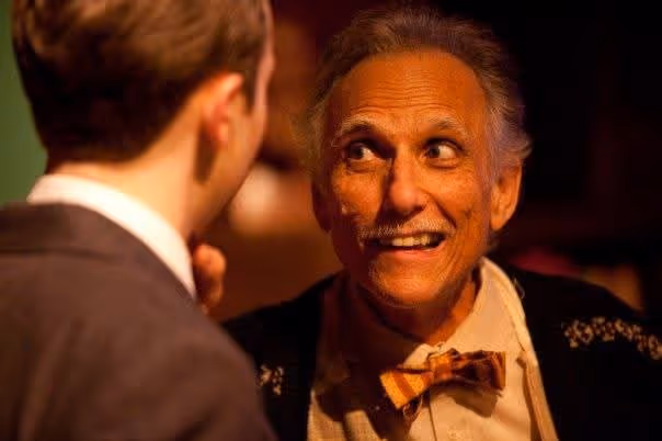 Older man with gray hair and a bow tie smiling and talking to a younger man in a suit.