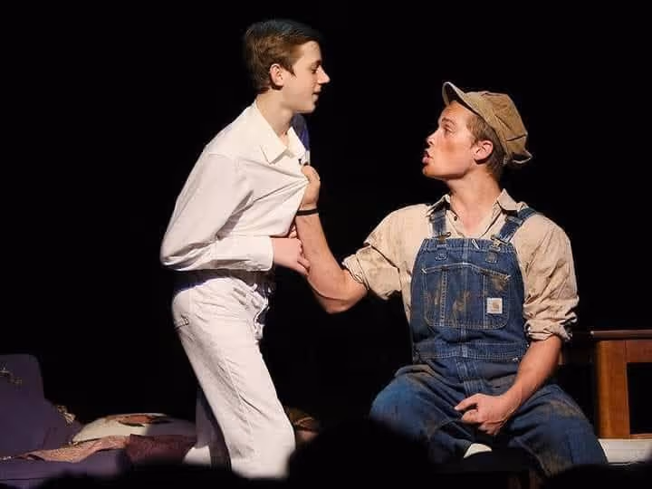 Two actors performing a tense scene on stage, one gripping the other's white shirt while sitting, the other standing and leaning forward.