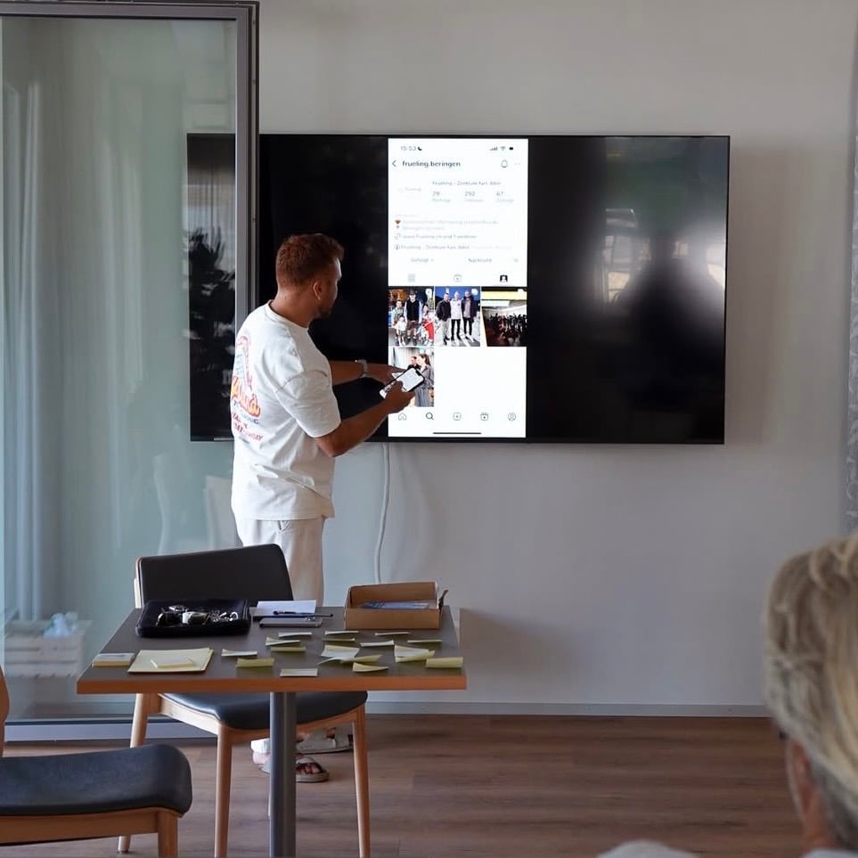 Man in white shirt and pants standing in front of a wall-mounted TV screen, projecting a phone's Instagram page, while gesturing with a smartphone in hand.