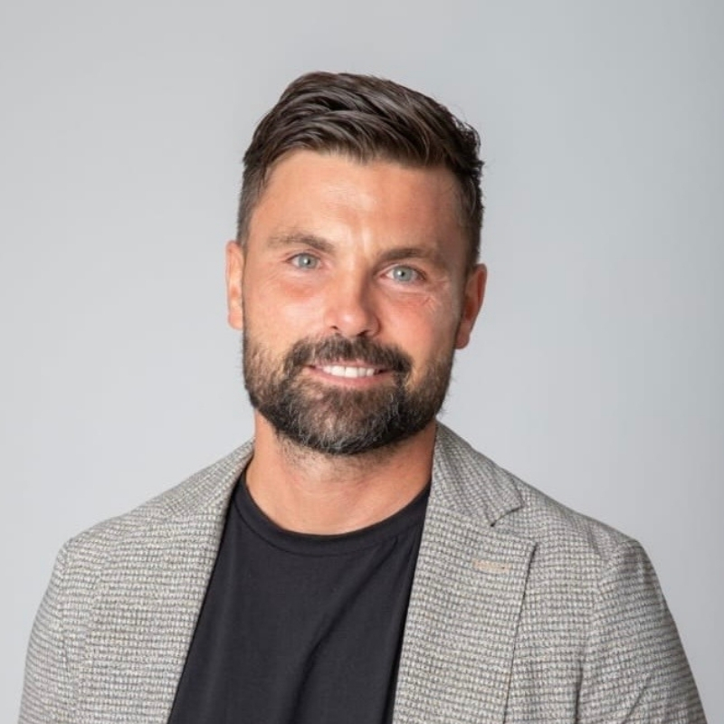Smiling man with dark hair and beard wearing a grey checkered blazer and black shirt against a plain light grey background.