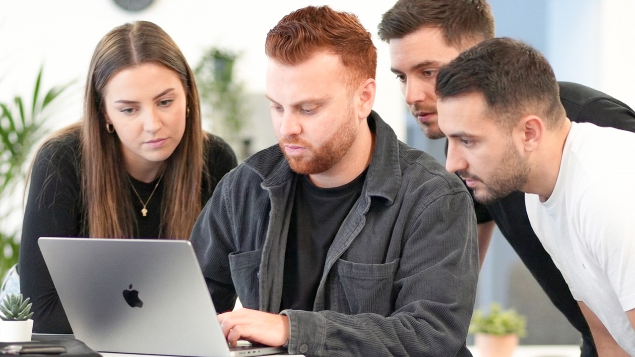 Four young adults focused on a laptop screen in a bright office with plants.