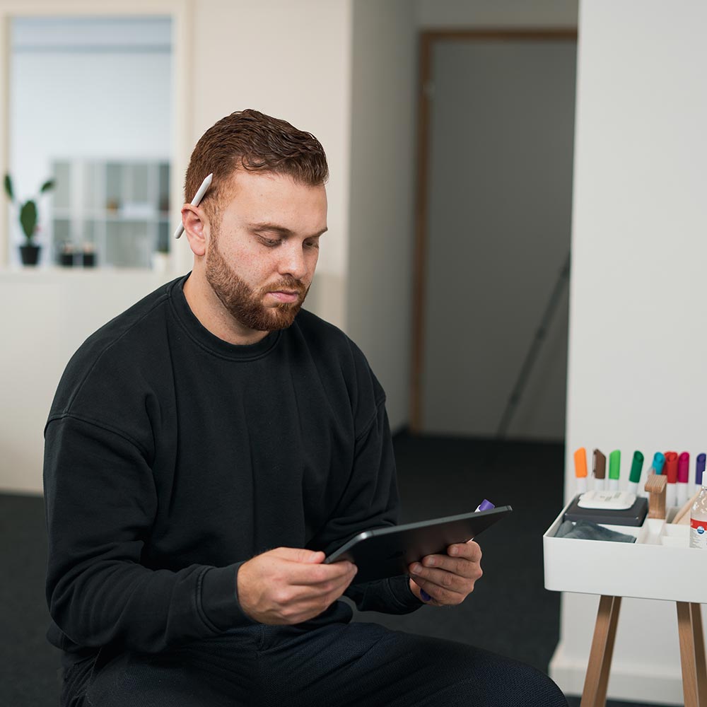 Man in a black sweatshirt holding a tablet and a purple marker, with a whiteboard and colorful markers in the background.