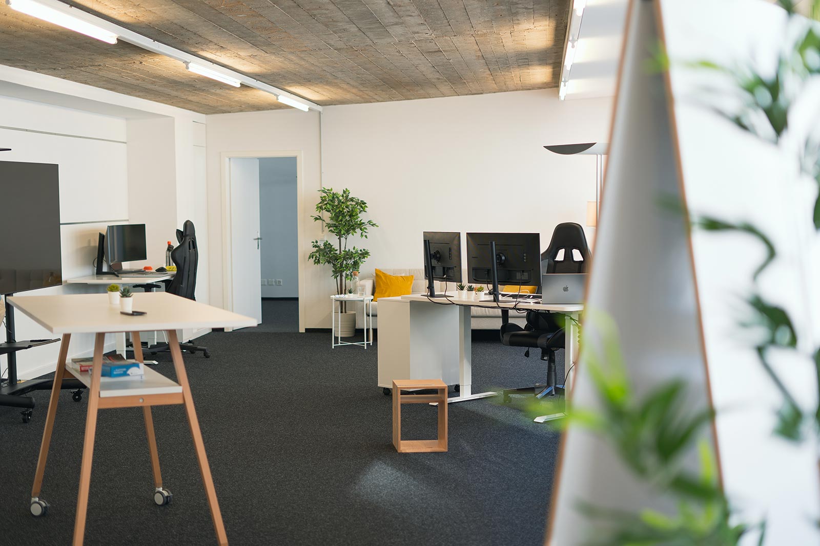 Modern office space with dual computer monitors on desks, black office chairs, a sofa with a yellow pillow, a potted plant, and a tall whiteboard on wheels.
