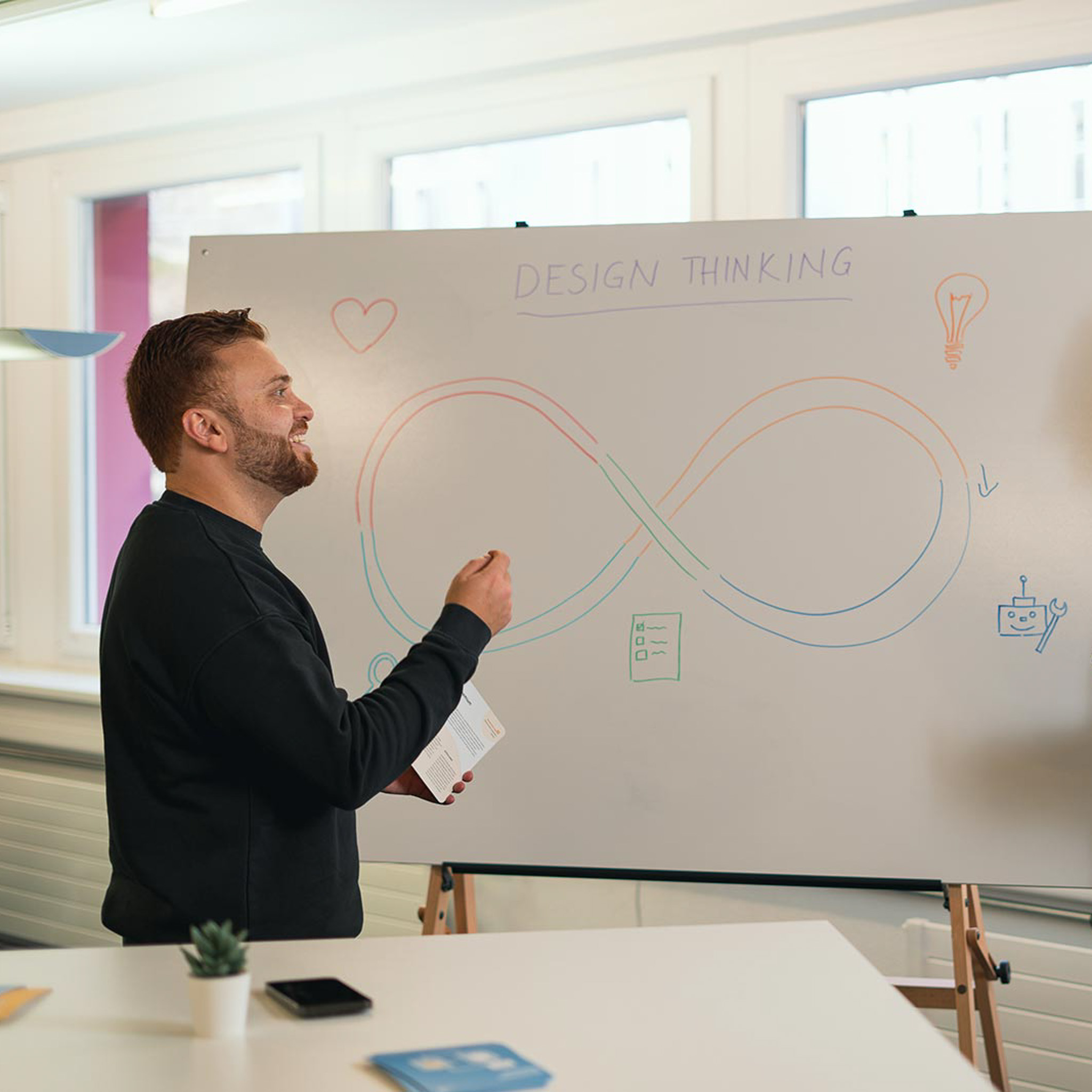 Man in black sweater smiling and presenting a design thinking diagram on a whiteboard with colorful infinity loop and icons.