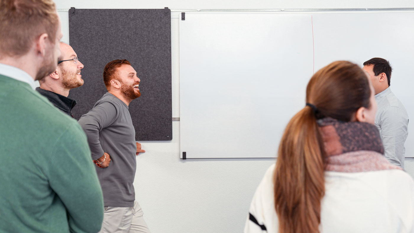 Group of five people in a casual meeting room standing and smiling near a whiteboard.