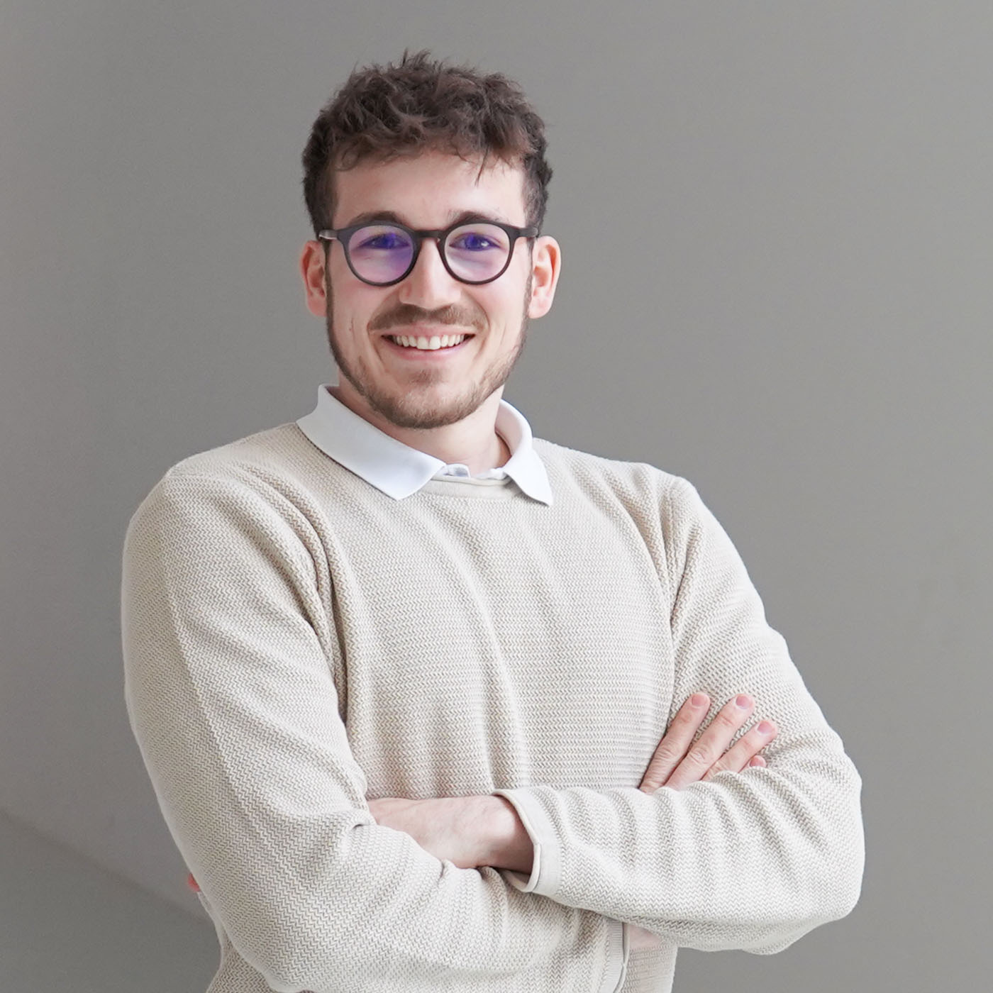 Smiling man with dark hair and beard wearing a grey checkered blazer and black shirt against a plain light grey background.