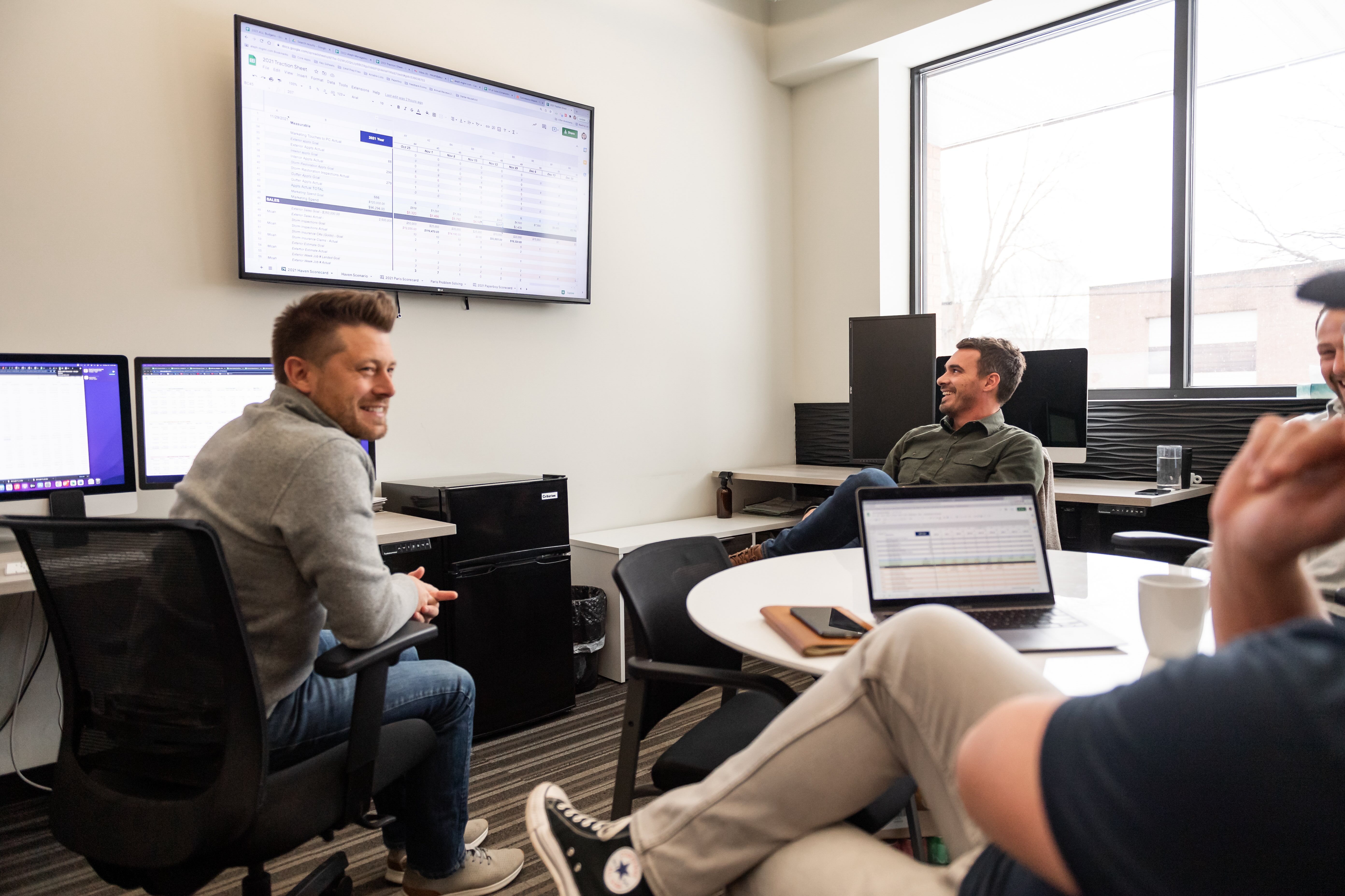 Three men in casual attire having a discussion in an office with laptops and a large screen displaying a spreadsheet.