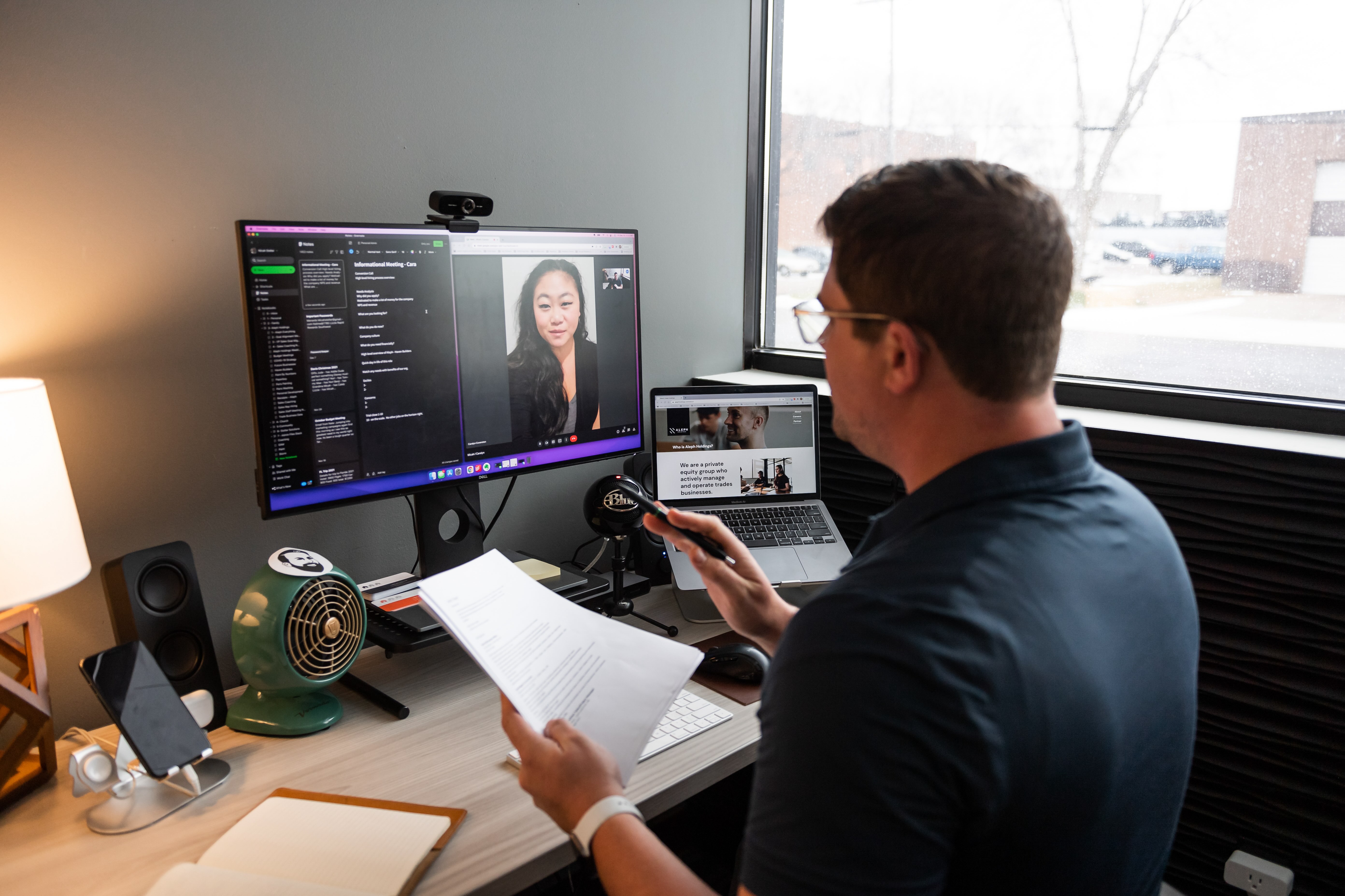 Man with glasses holding papers, participating in a video call at a desk with two monitors, a phone, and a lamp near a window.
