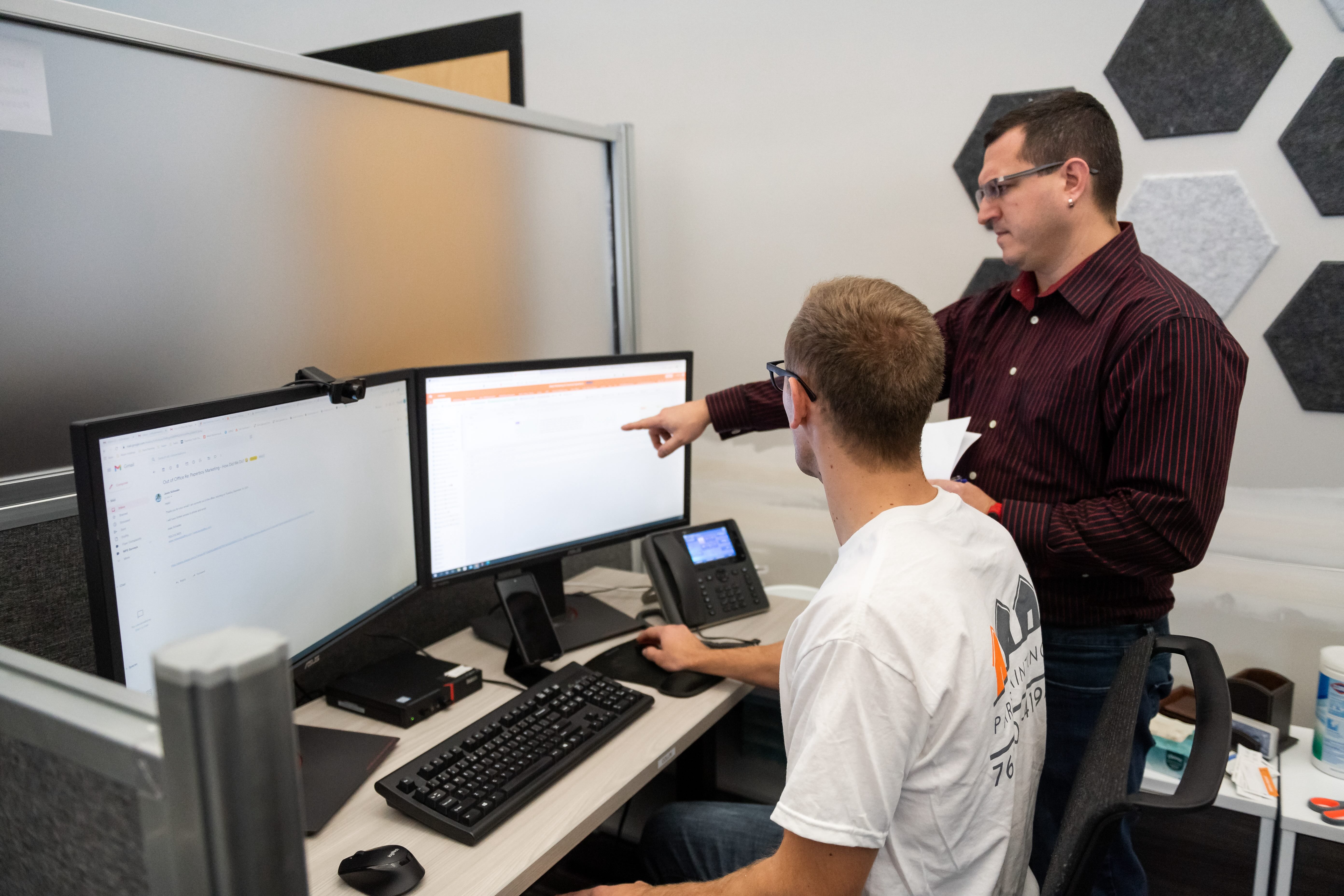 Two men working together at a desk with dual monitors, one seated and the other standing and pointing at one screen.