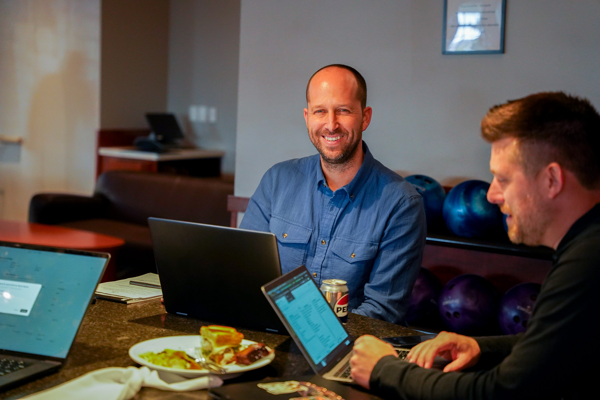 Man with glasses holding papers, participating in a video call at a desk with two monitors, a phone, and a lamp near a window.