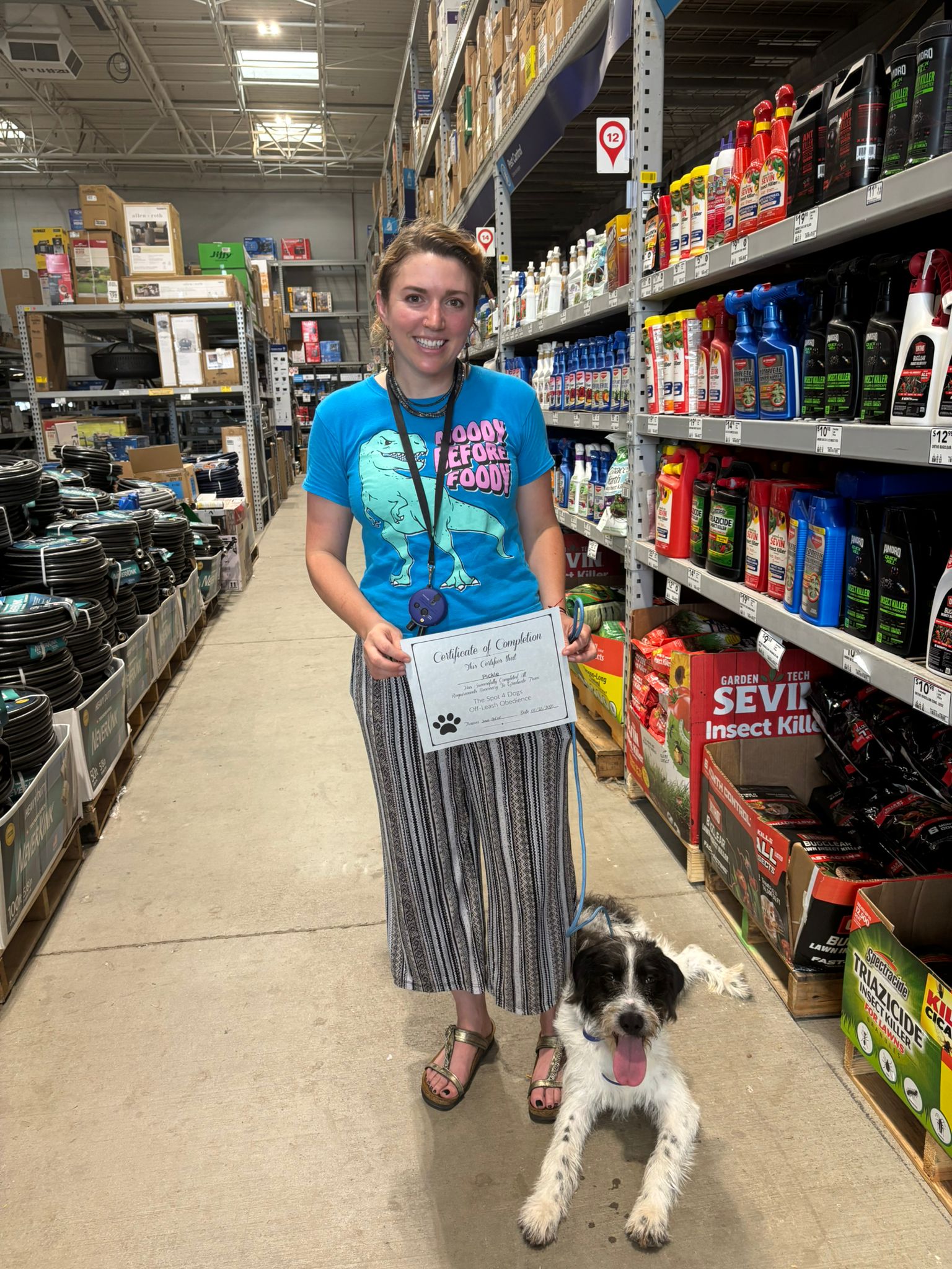 Smiling woman in a blue dinosaur shirt holding a certificate standing in a store aisle with a black and white dog lying beside her.