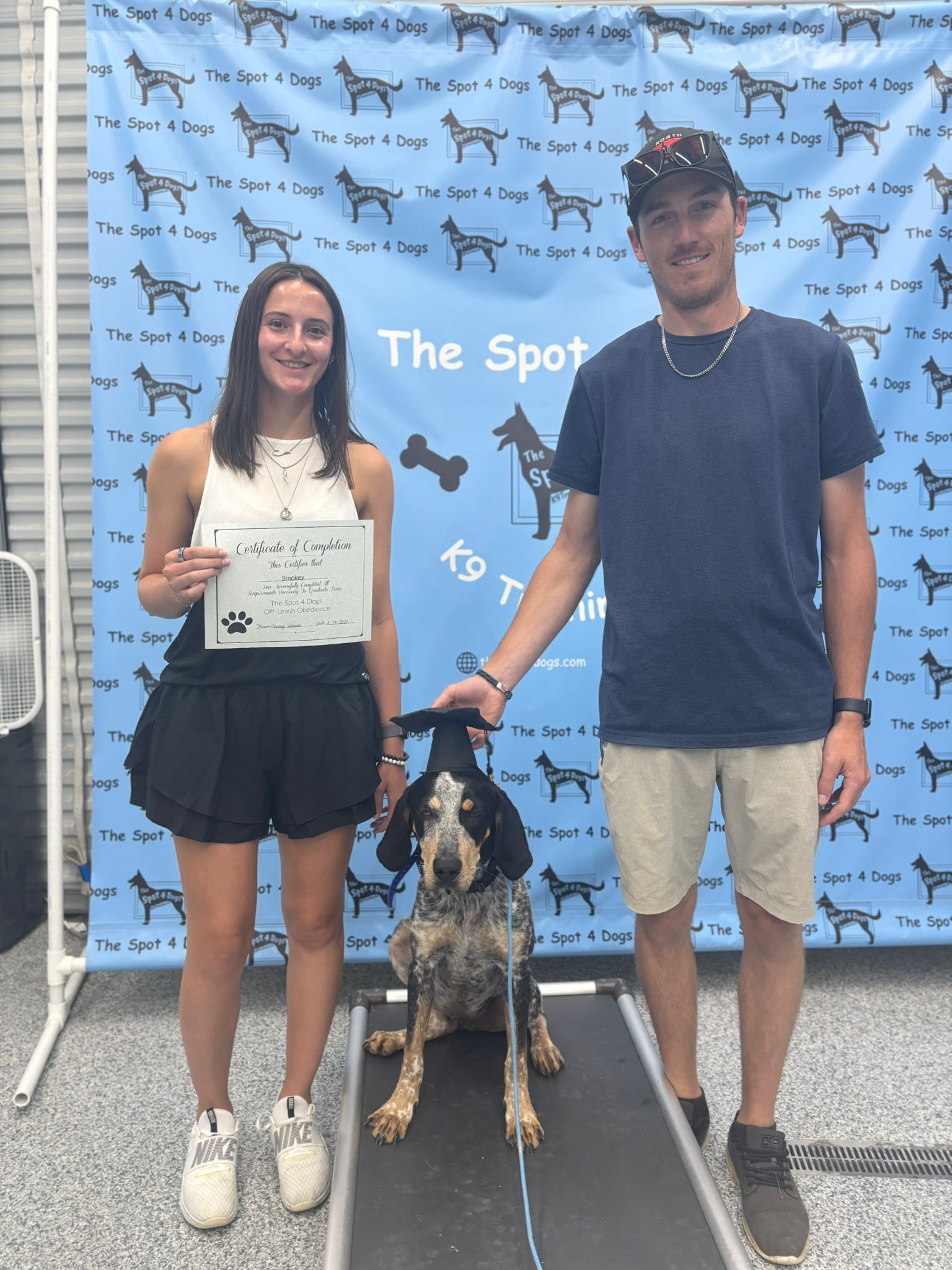 Woman holding certificate beside man and dog wearing graduation cap in front of The Spot 4 Dogs banner.