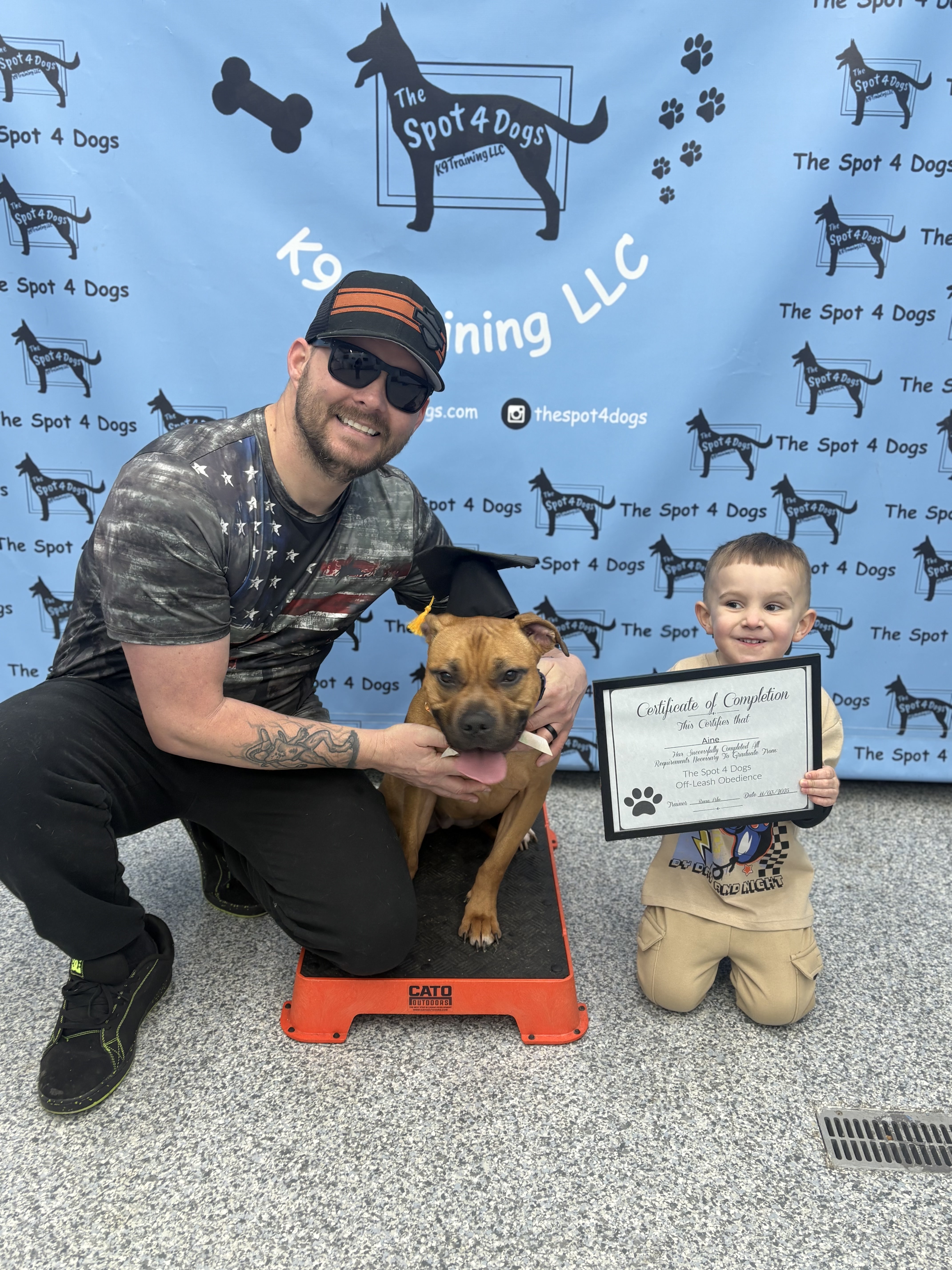 Man and young boy posing with a brown dog wearing a graduation cap, the boy holding a certificate of completion from The Spot 4 Dogs dog training.