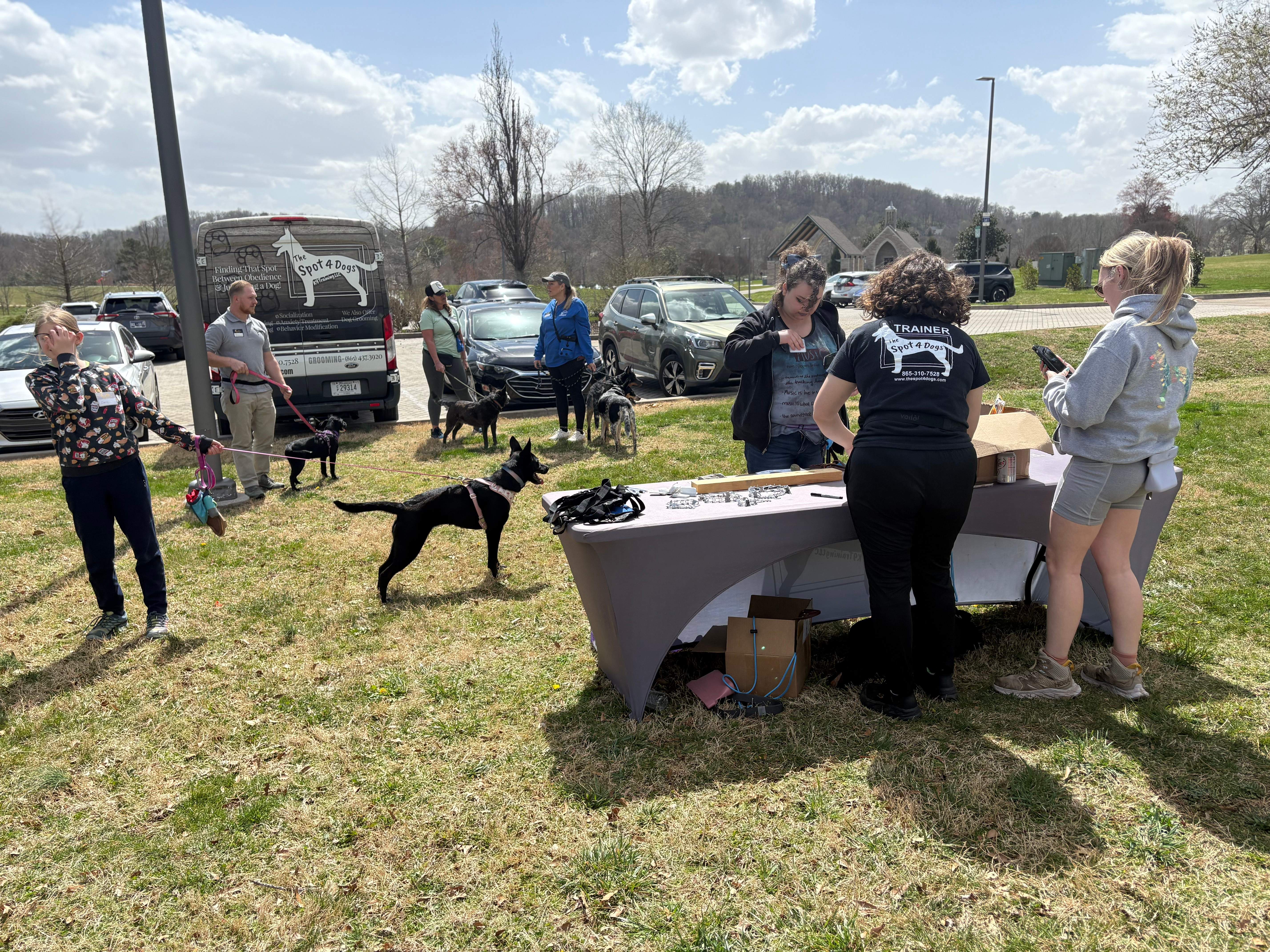 Several people with dogs gathered outdoors near a table with supplies, in front of a van labeled Spot 4 Dogs.