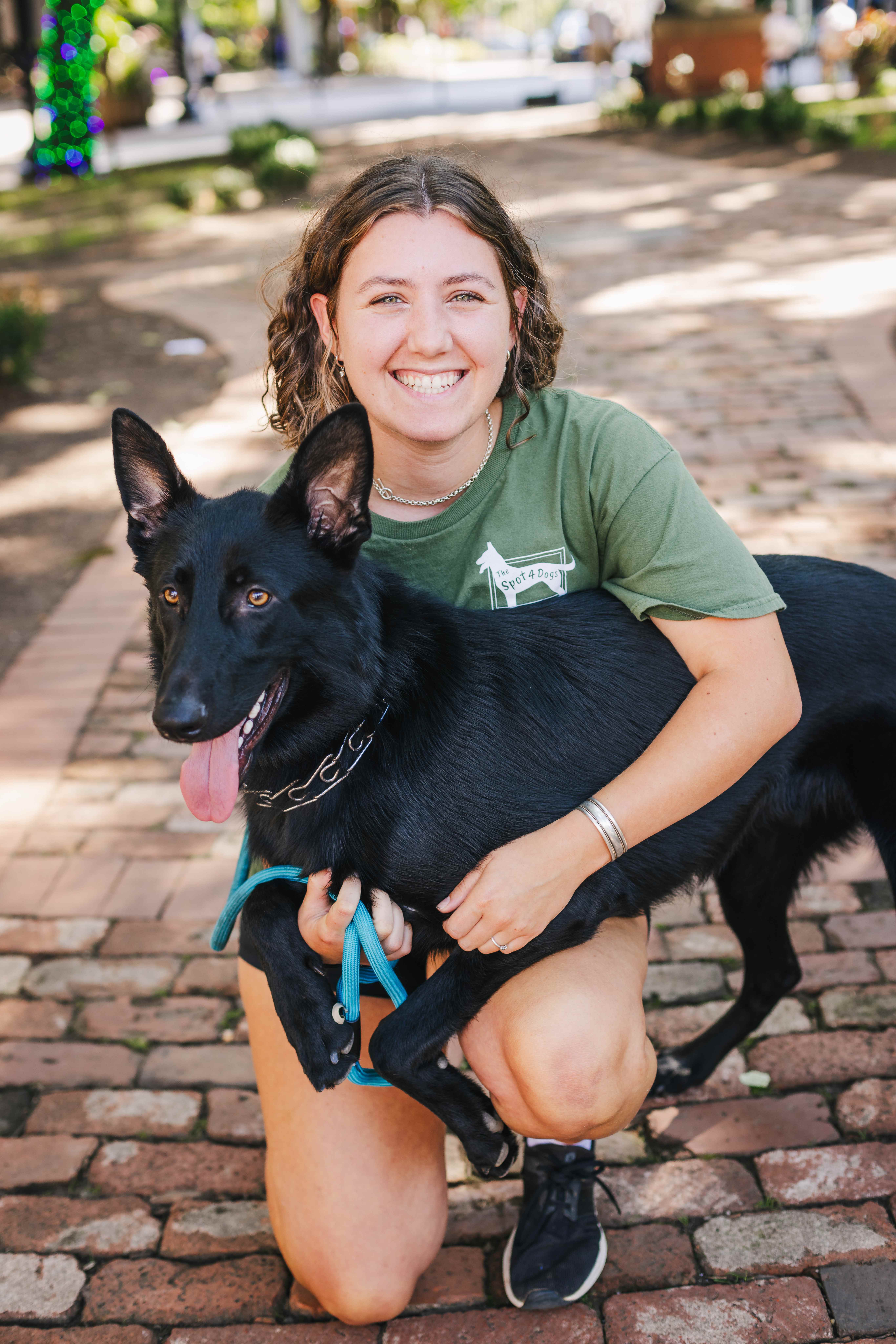 Smiling young woman kneeling on a brick path holding a happy black German Shepherd with a blue leash.