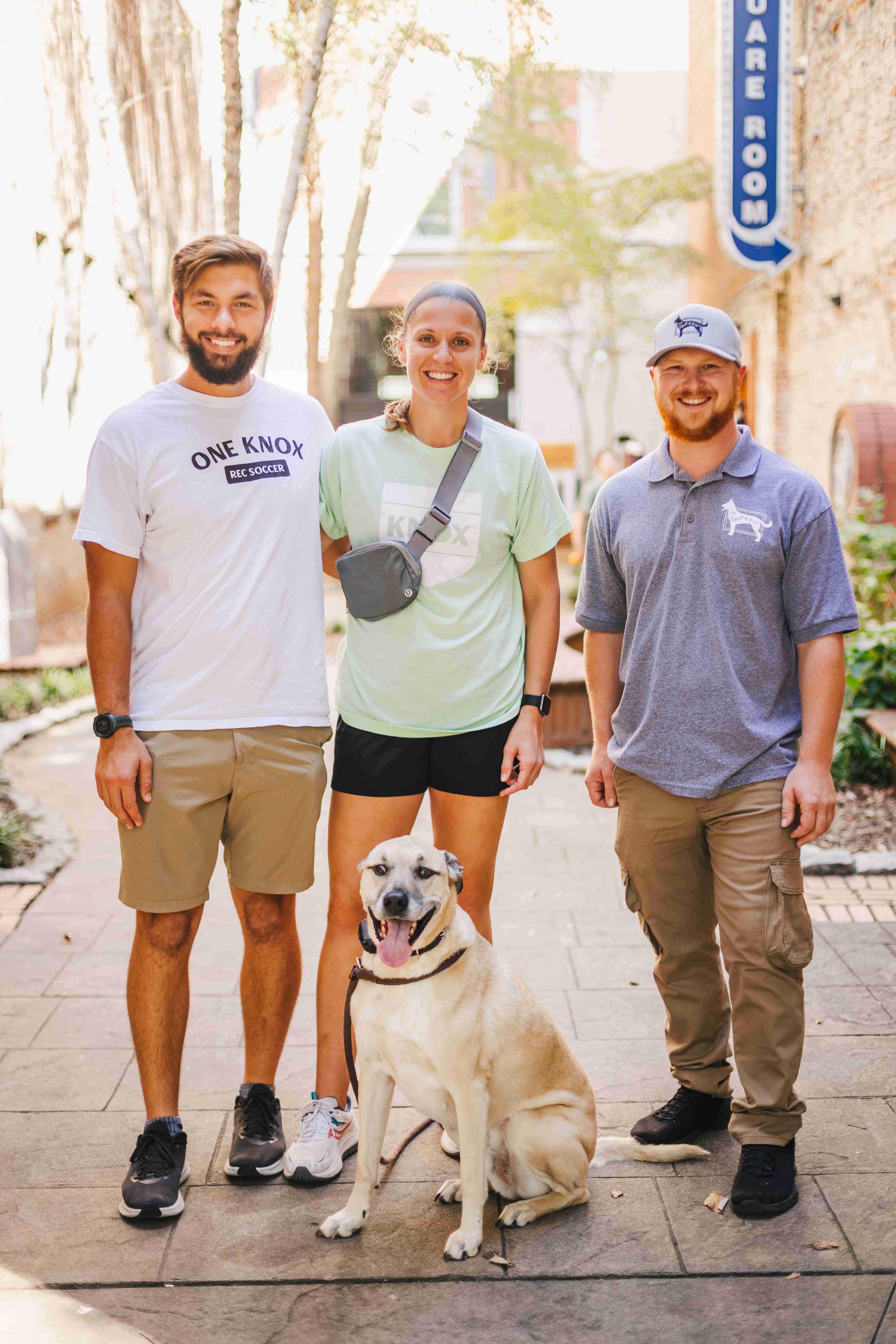 Three smiling adults with a happy tan dog sitting in front of them on a paved outdoor path.