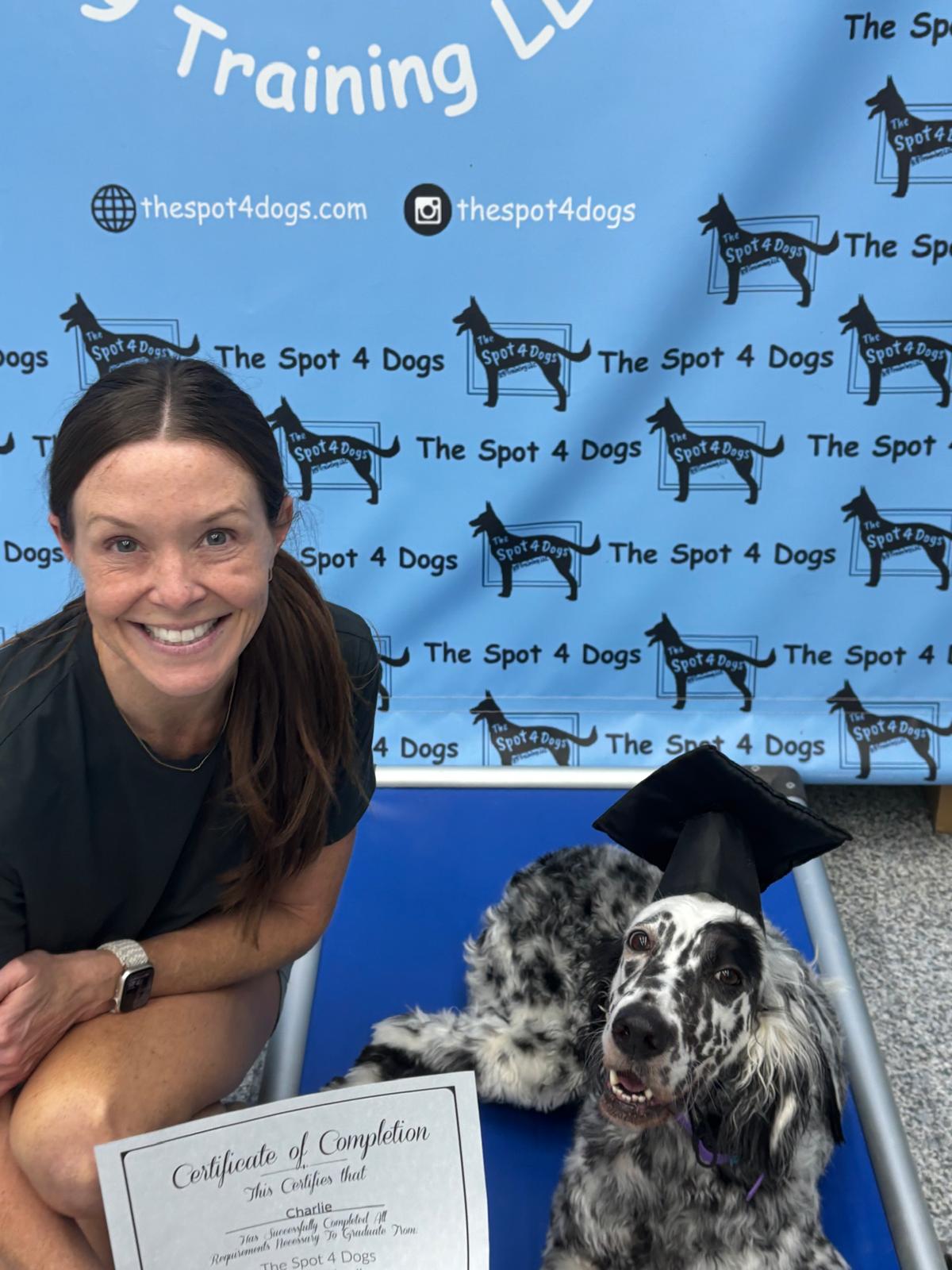 Smiling woman kneels next to a black and white dog wearing a graduation cap, holding a certificate of completion from The Spot 4 Dogs.