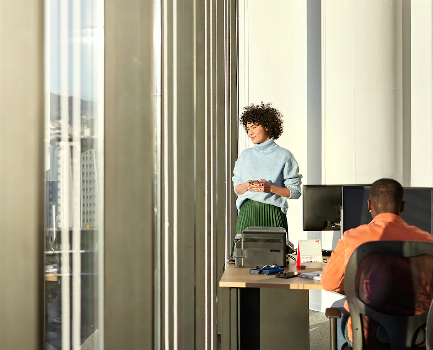 Woman in a blue sweater standing by a window looking at her phone while a man in an orange shirt works at a desk with computers.