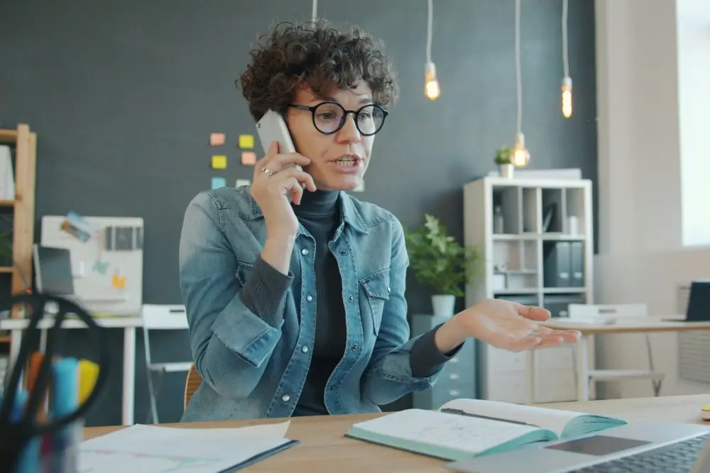 A woman with curly hair wearing a denim jacket, holding a phone to her ear with one hand raised in a questioning gesture, in a home or office setting.