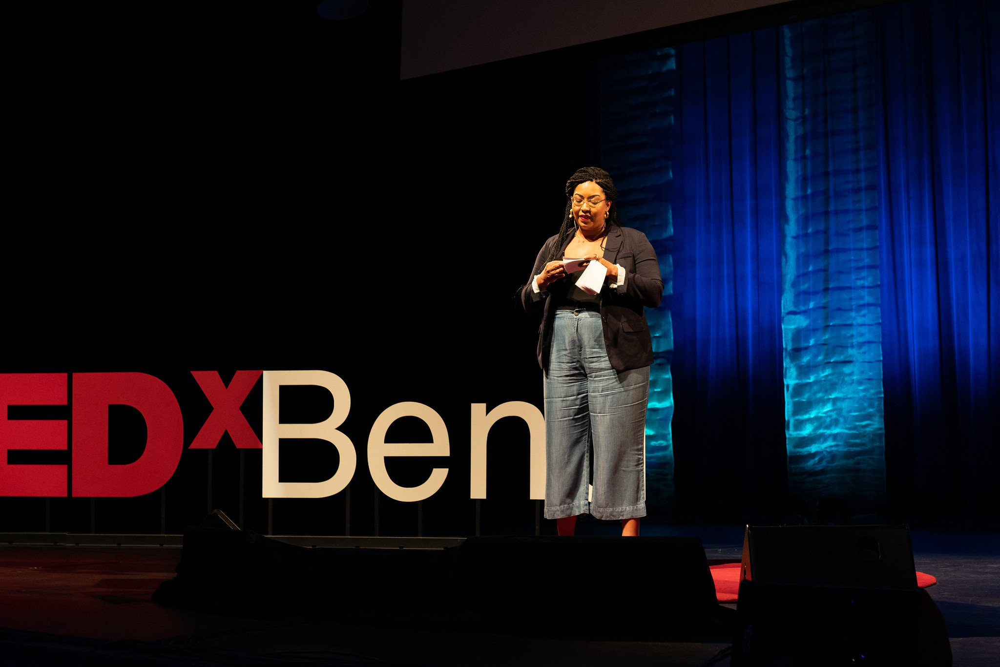 Speaker with glasses and braided hair holding notes on a TEDx stage with blue curtains in the background.
