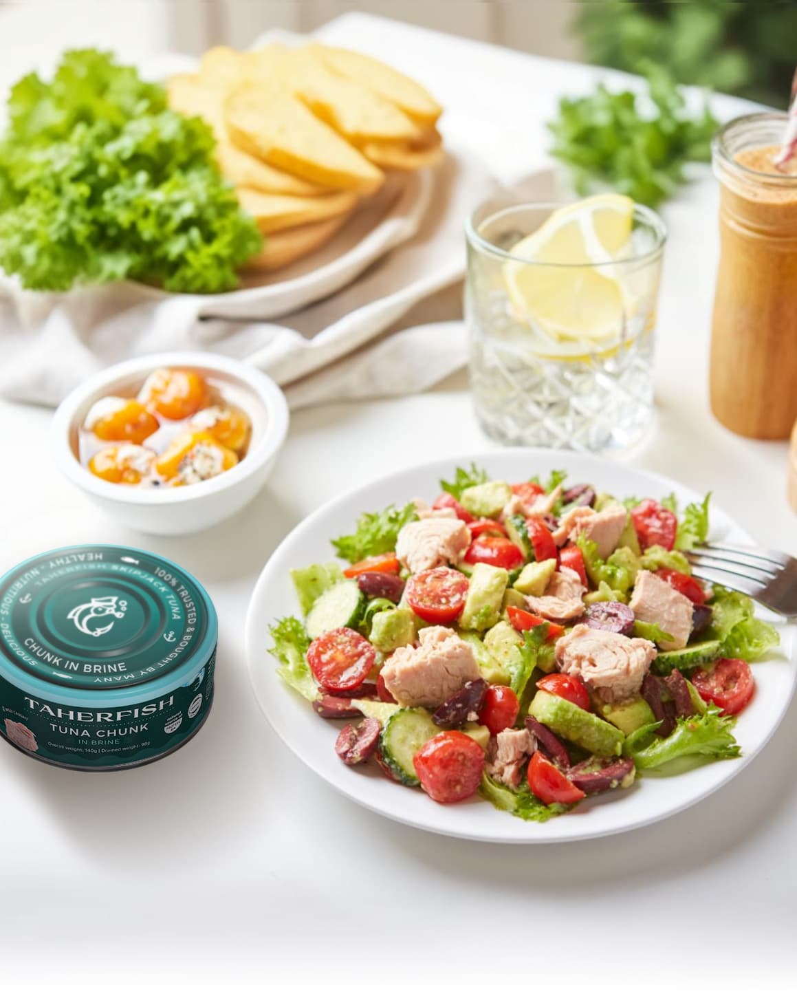 Plate of fresh salad with tuna chunks, cherry tomatoes, cucumbers, olives, and lettuce with a fork on the side, accompanied by a glass of water with lemon, a jar of dressing, bread slices, and a can of Taherfish tuna chunk.