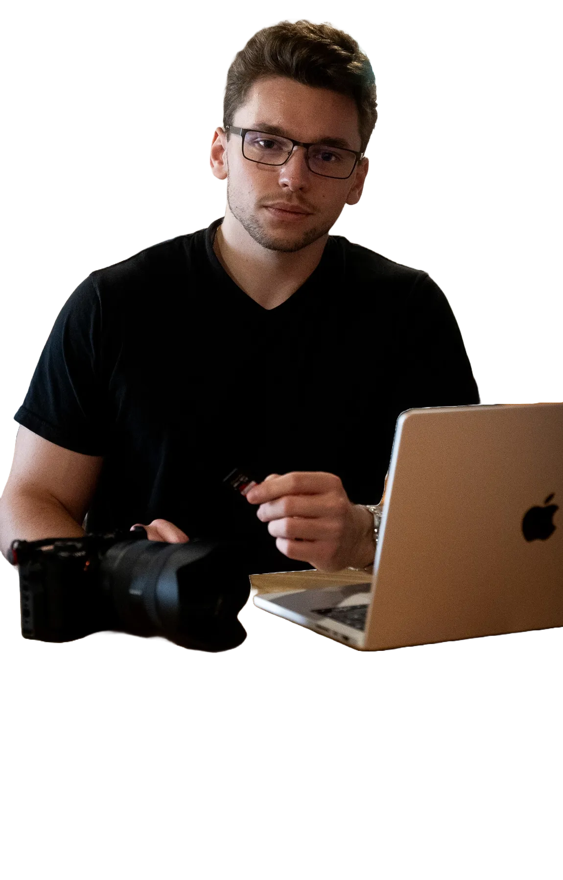 Man wearing glasses and a black t-shirt, holding a memory card next to a camera and an open laptop.