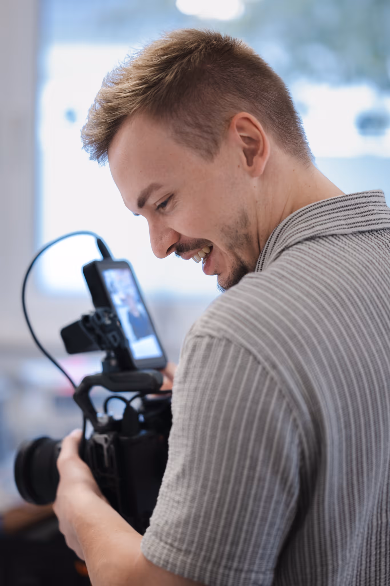 Smiling man adjusting a professional camera with an attached monitor in an indoor setting.