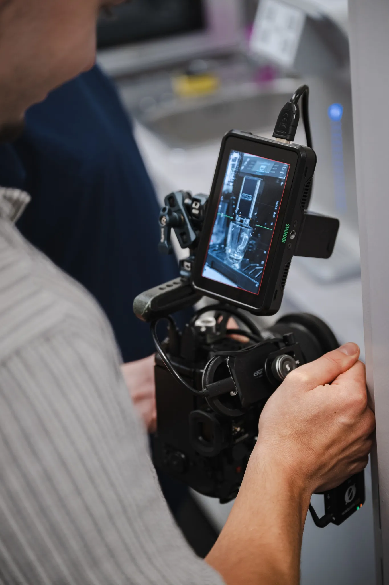 Person operating a professional video camera with a monitor displaying a close-up shot of a coffee machine pouring coffee into a glass.