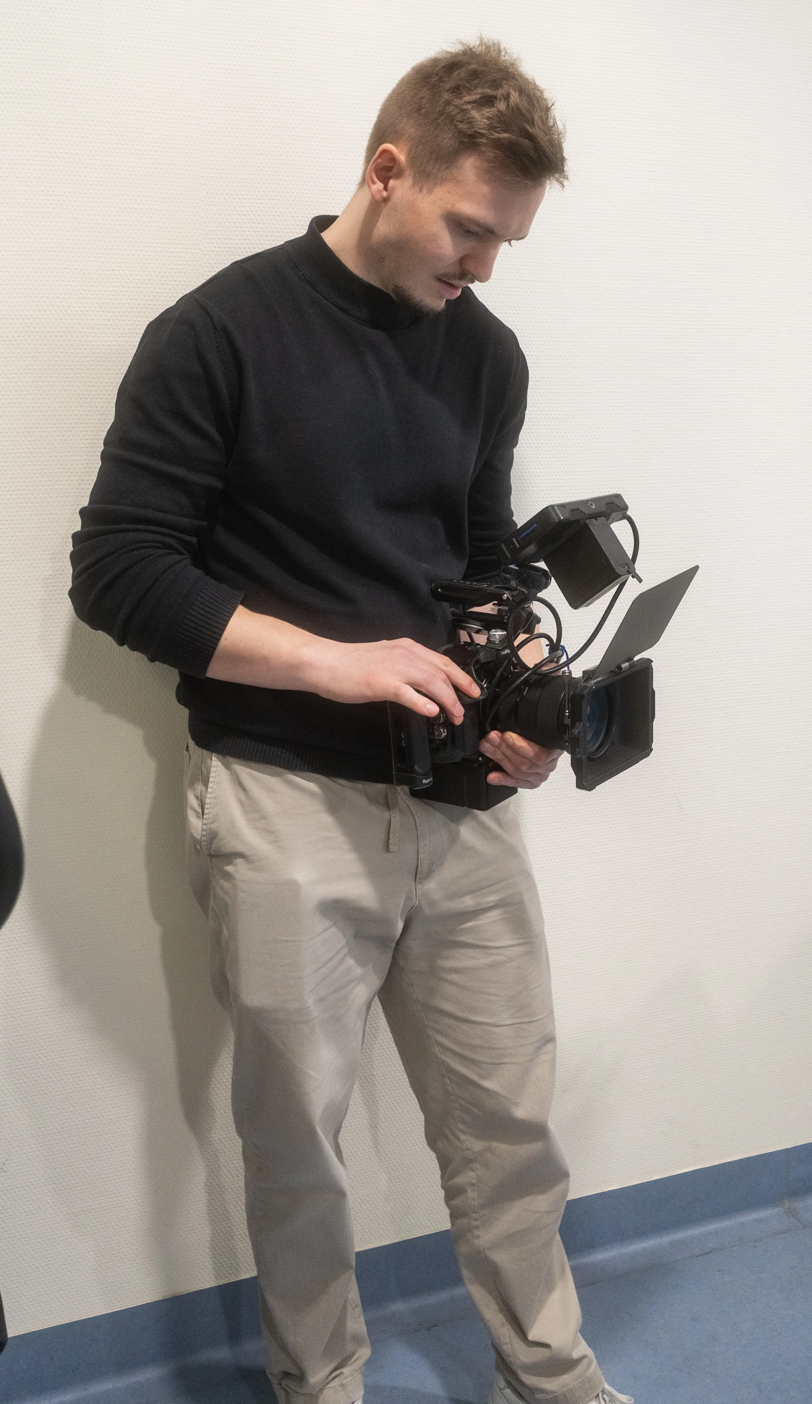Man wearing black sweater and beige pants operating a professional video camera indoors against a white wall.