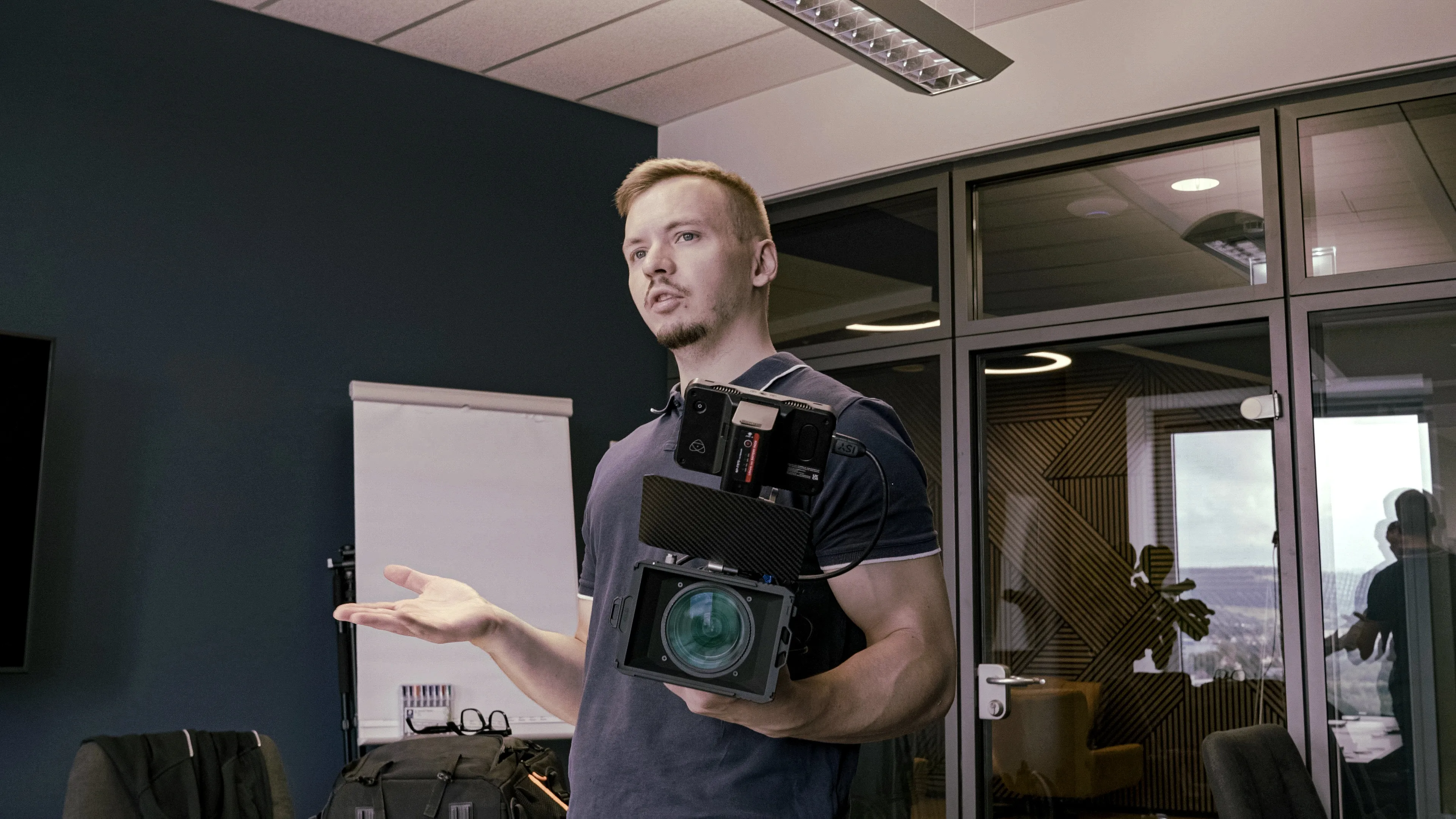 Man holding a professional video camera, gesturing with his other hand, standing in an office with glass walls and a flip chart in the background.