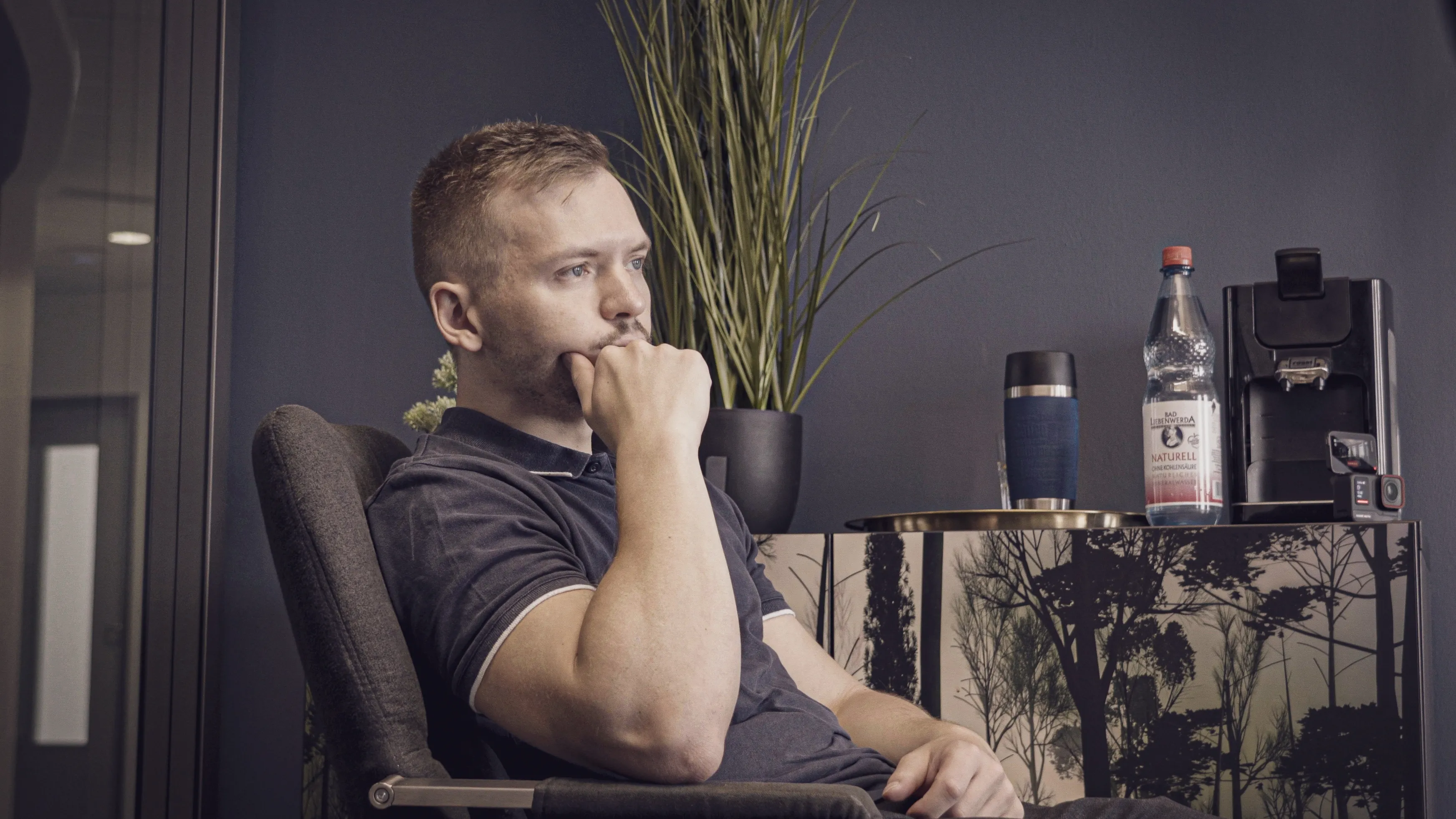 Man wearing black polo shirt sitting in an office chair, resting his chin on his hand and looking thoughtful.