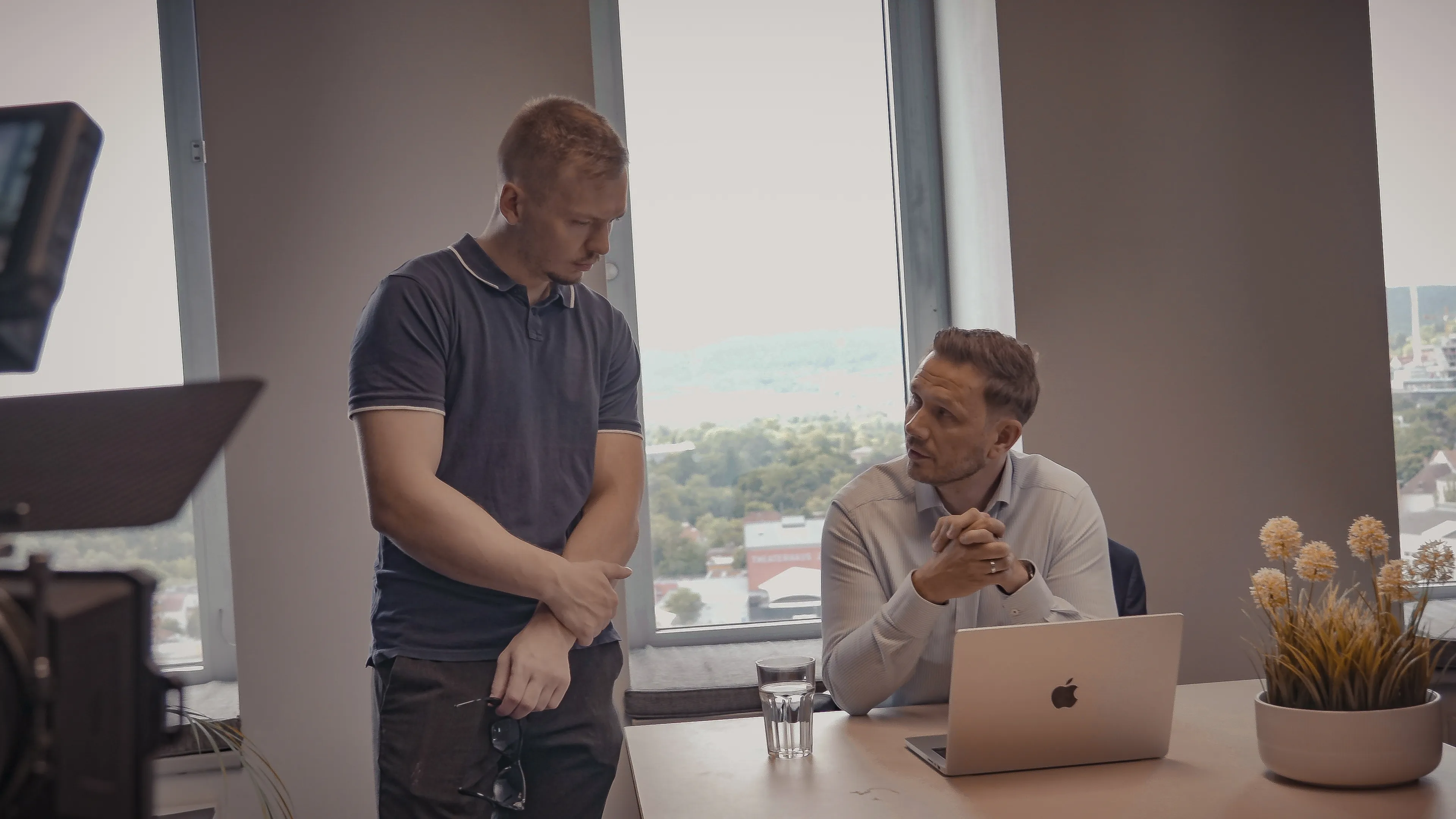 Two men in an office conversing, one standing with sunglasses in hand and the other seated at a desk with a laptop and a glass of water.