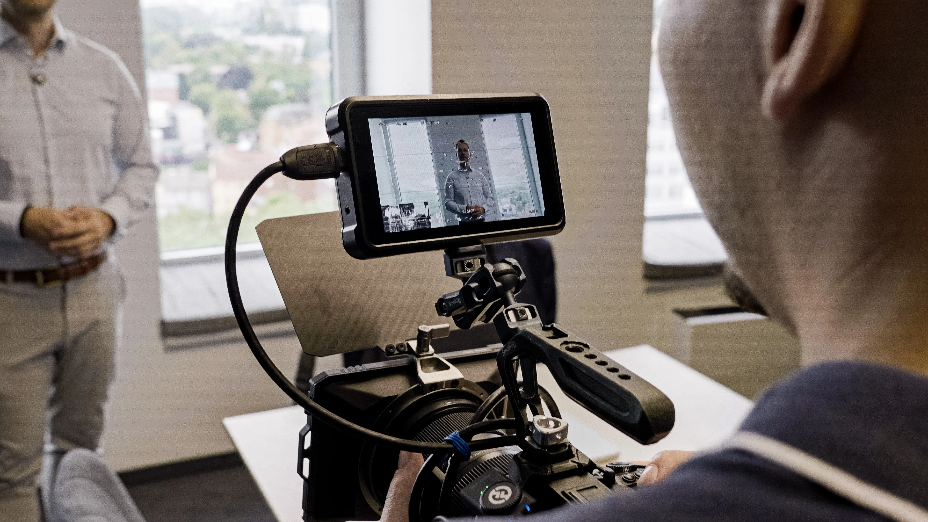 Person filming a man standing and speaking in an office with a digital camera's screen showing the man.