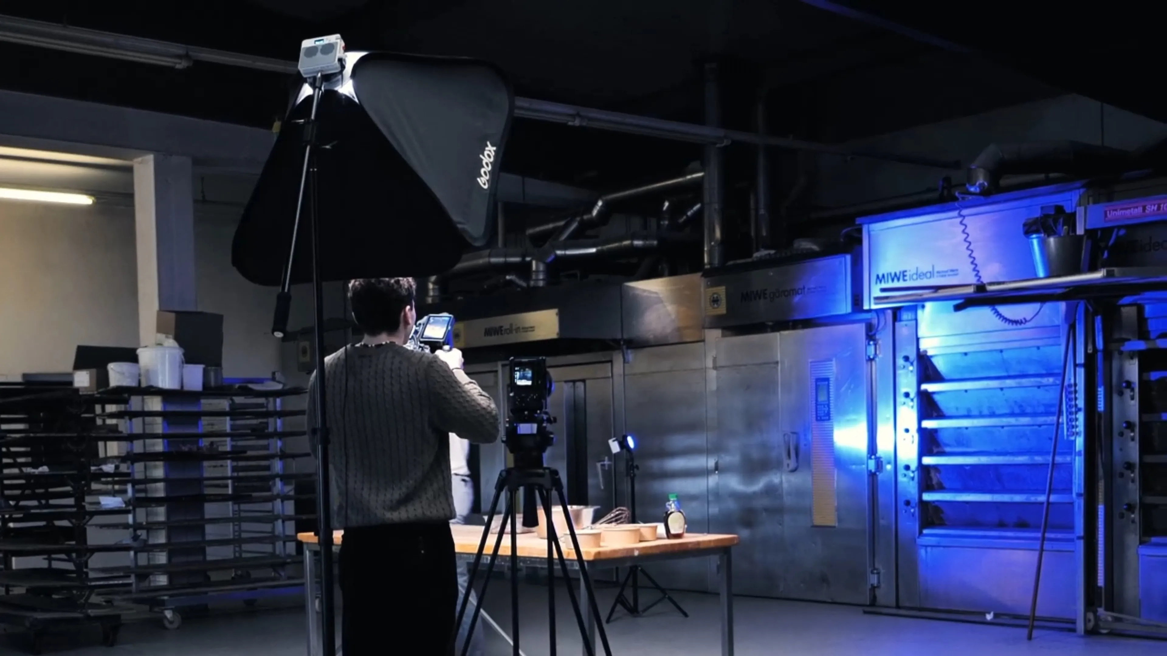 Person filming a table with baking items in an industrial kitchen studio with large ovens and a softbox light.