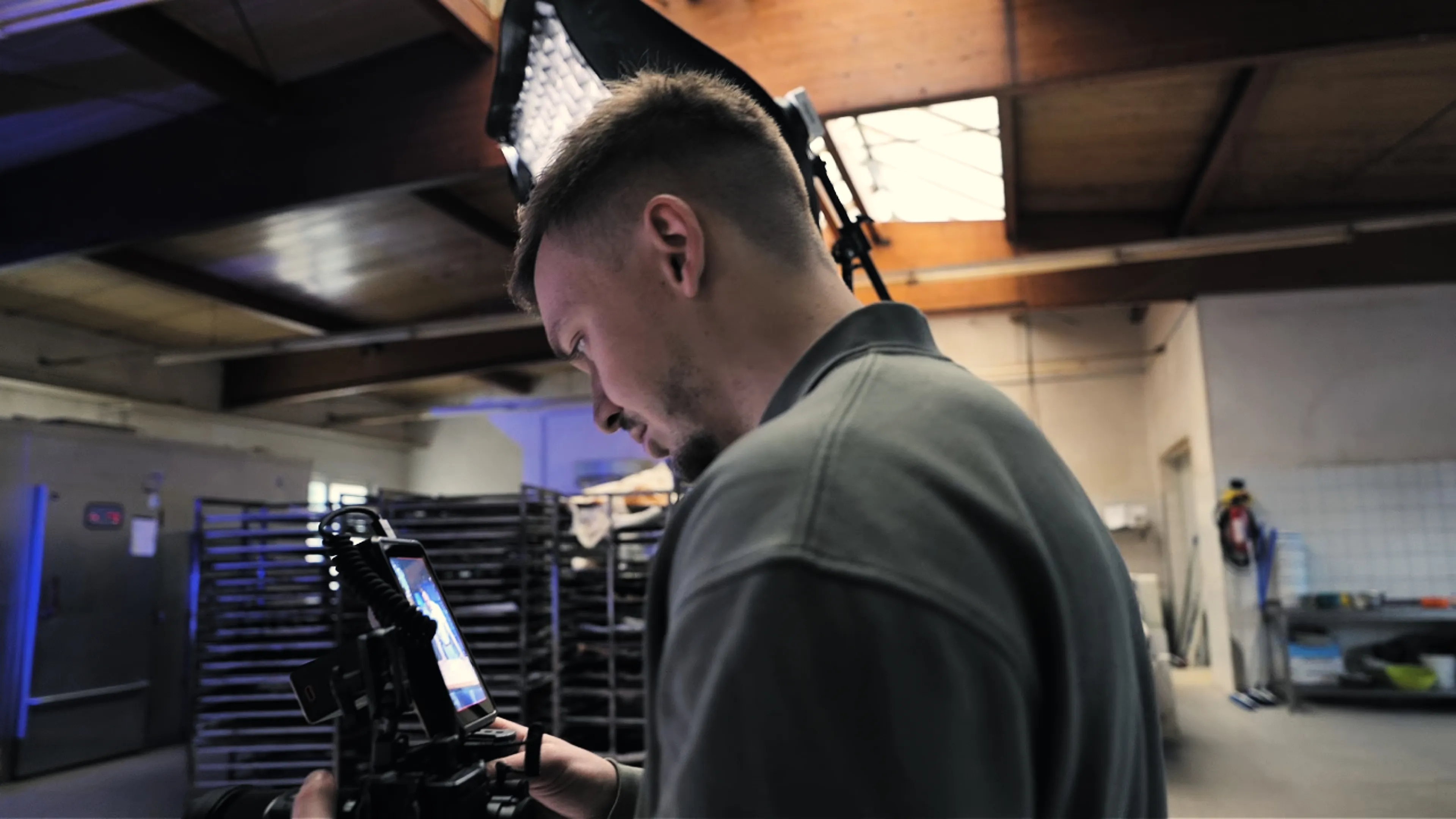 Man operating a professional video camera indoors with studio lighting and storage racks in the background.