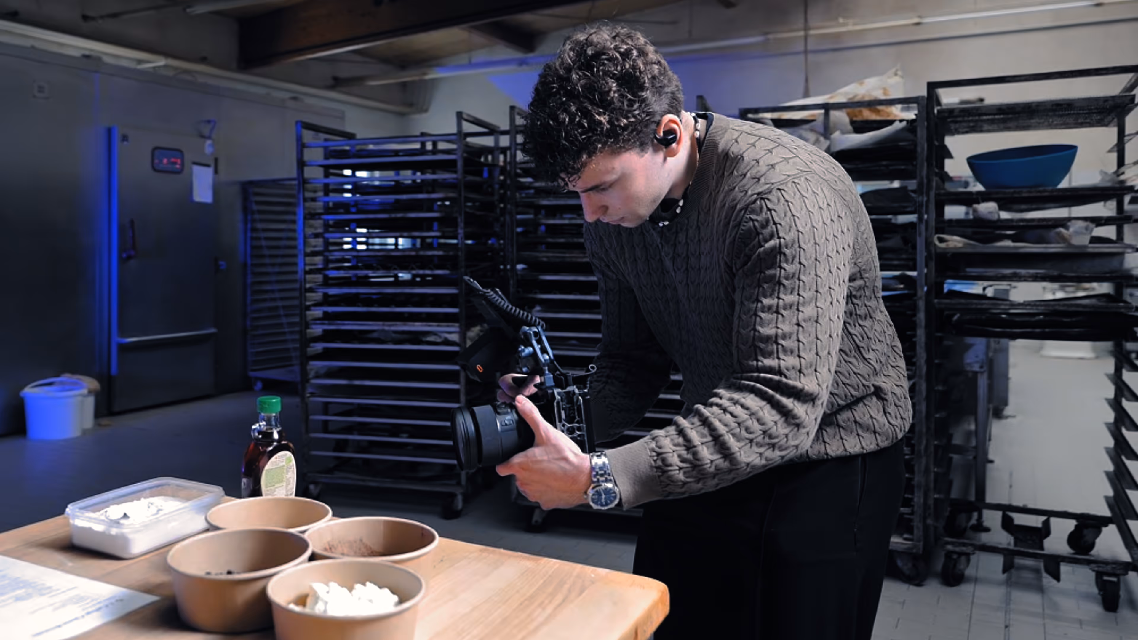 Man in a bakery kitchen filming ingredients on a wooden table with a professional camera.