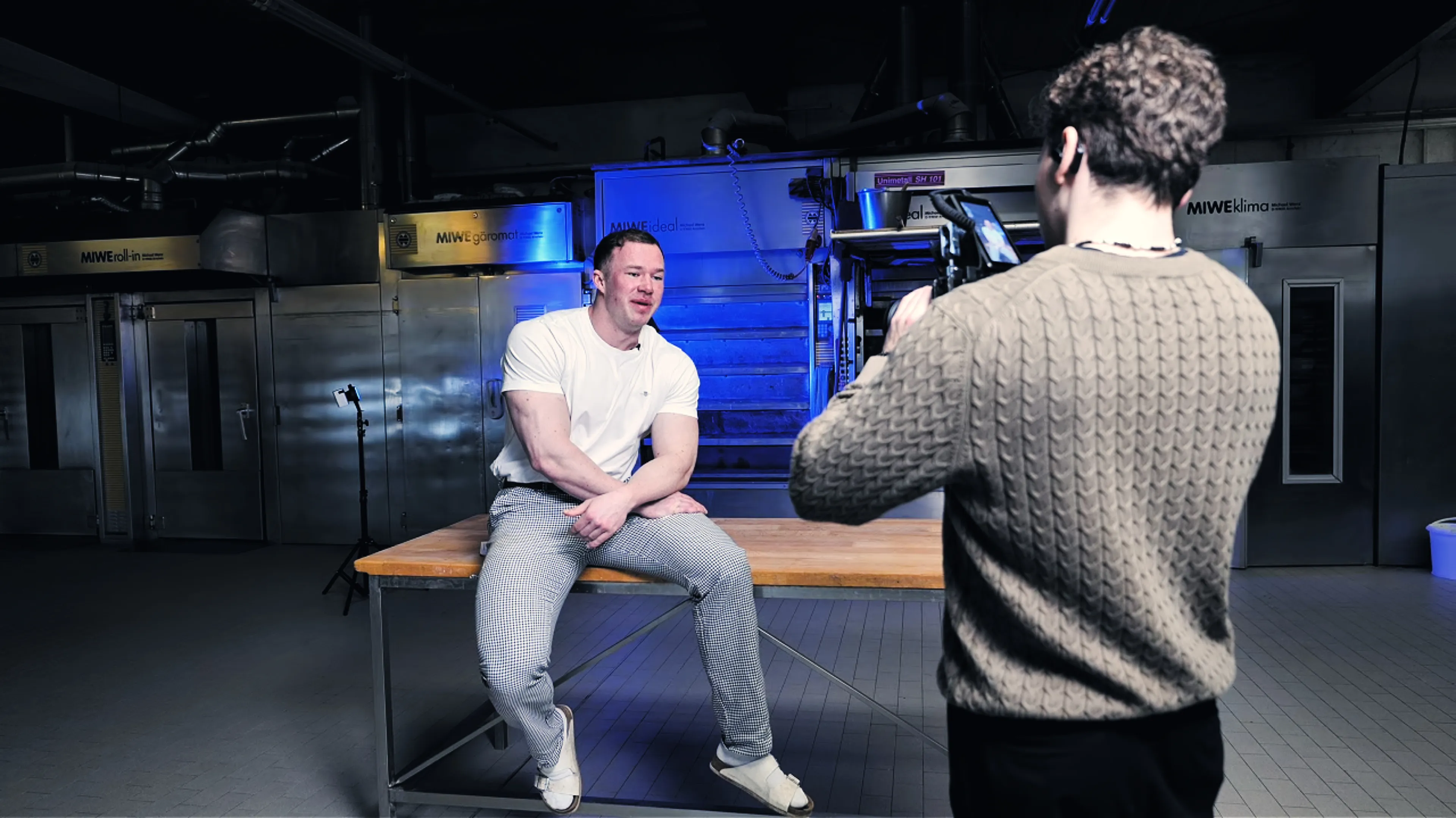 Man in white t-shirt and checkered pants sitting on a wooden table being filmed by another person in a brown sweater inside an industrial kitchen.