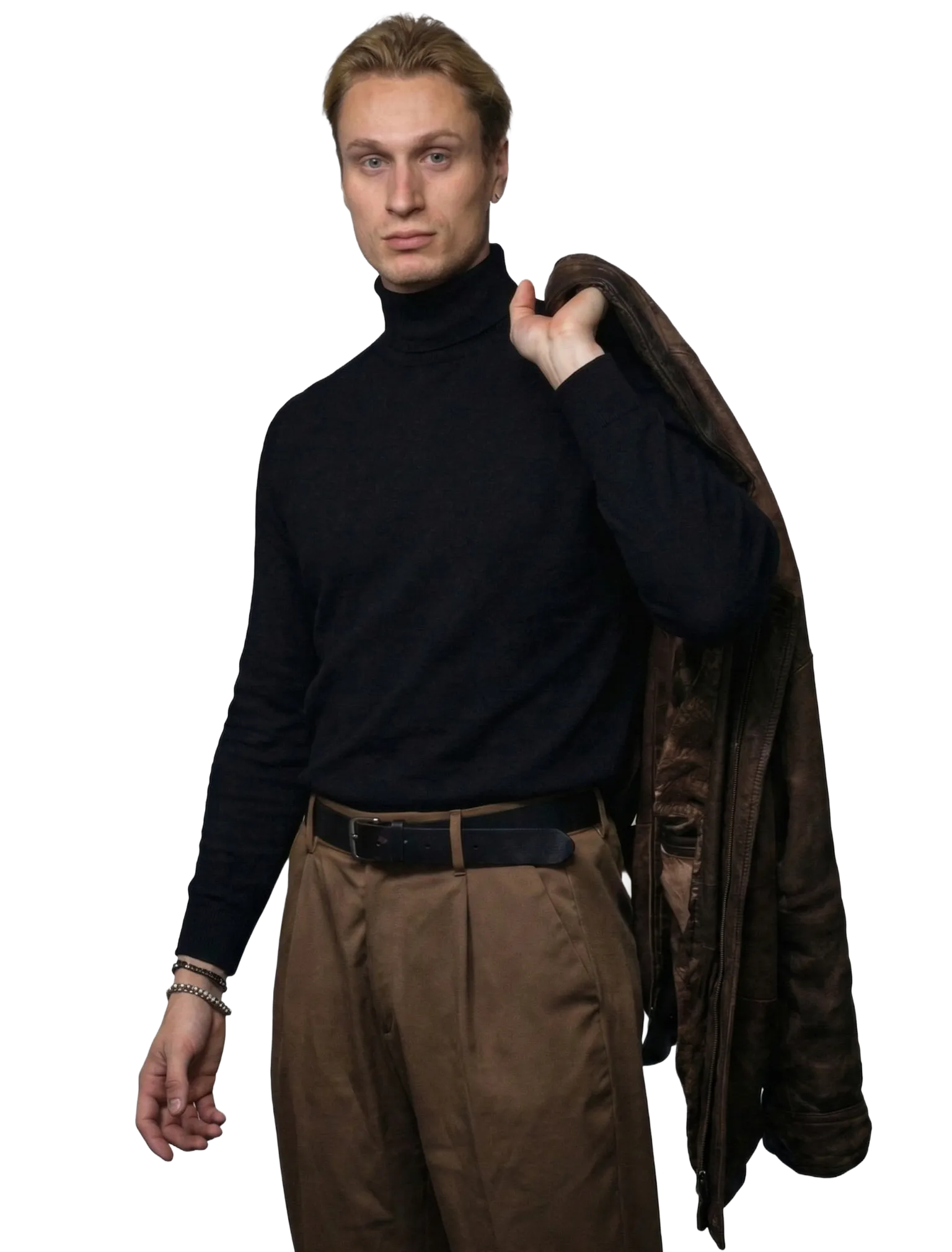 Blond man wearing a black turtleneck and brown pants holding a brown jacket over his shoulder.