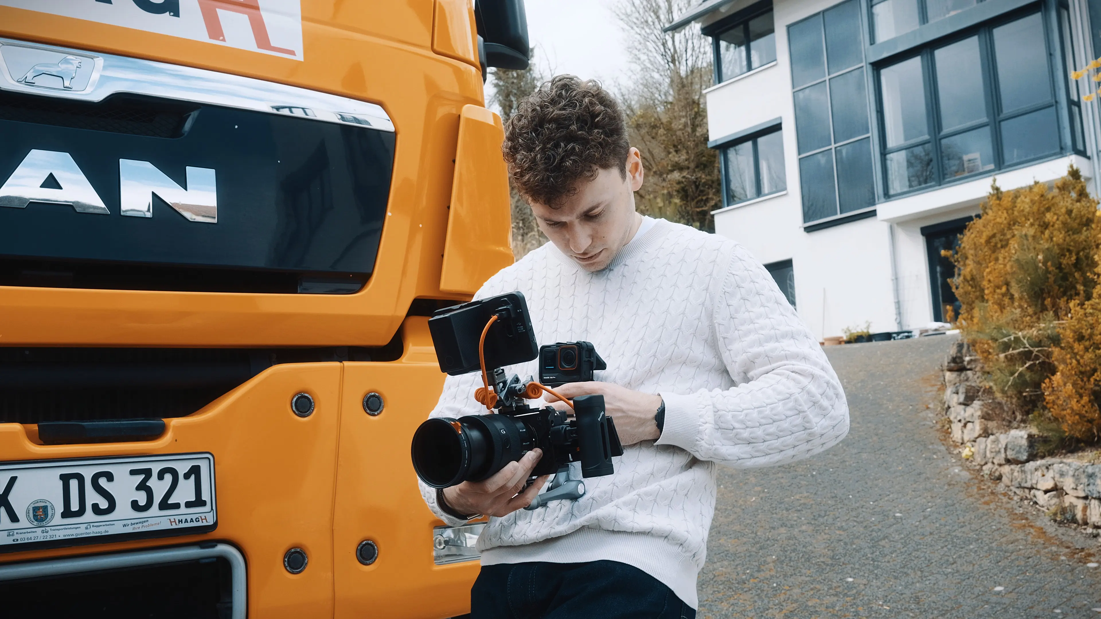 Man in white t-shirt and checkered pants sitting on a wooden table being filmed by another person in a brown sweater inside an industrial kitchen.