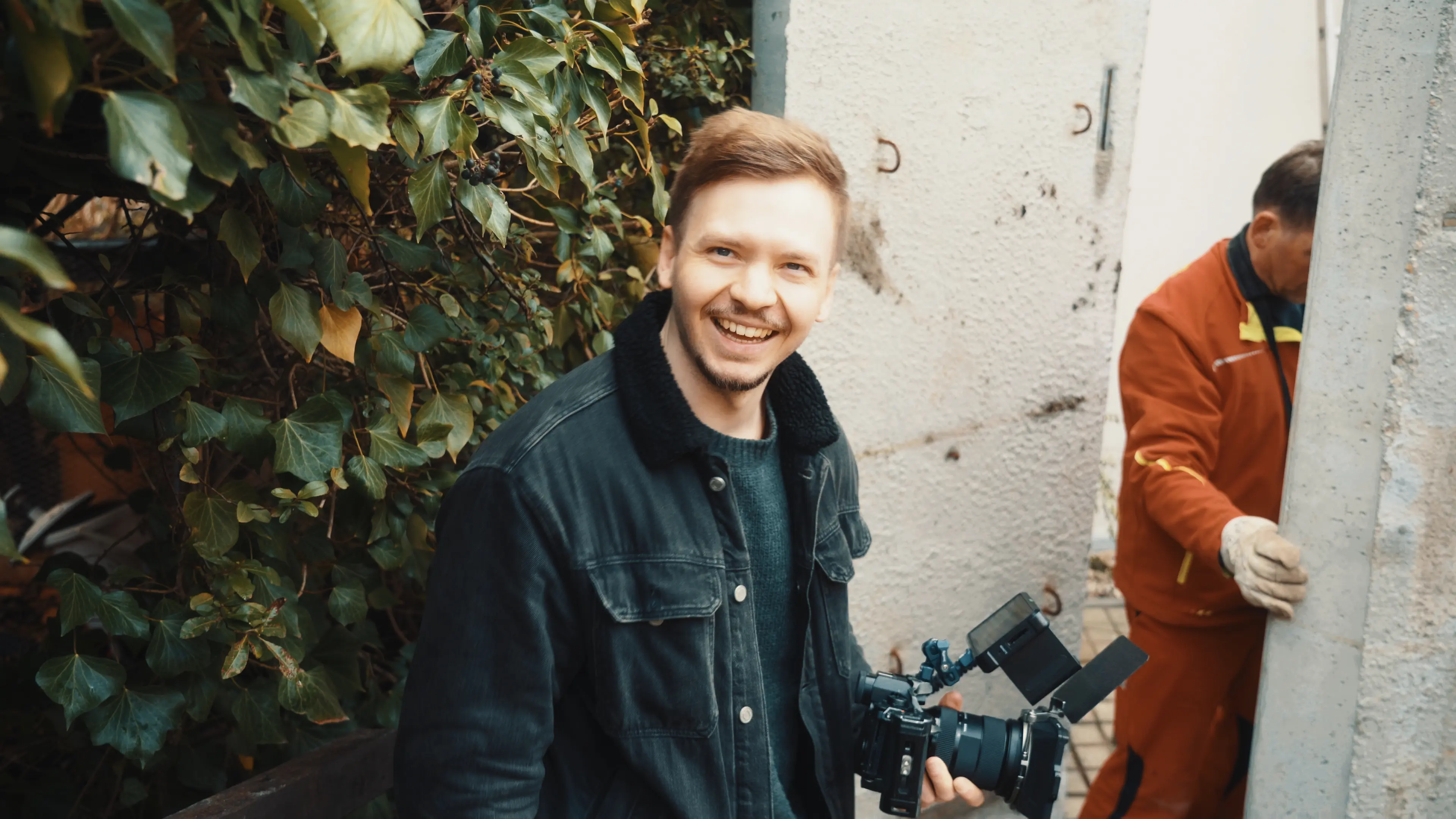 A young woman in a blue hoodie and beige pants walking on a city street with a man behind her holding a video camera.