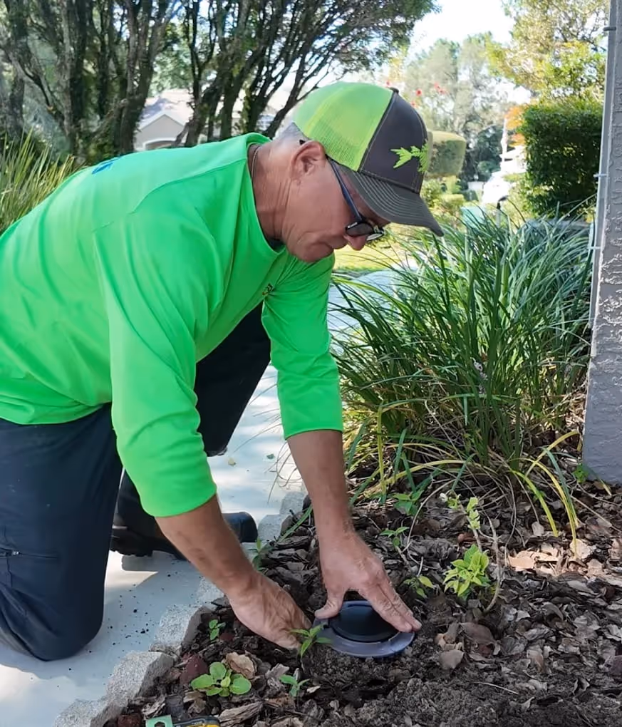 Man wearing a bright green shirt and cap installing a small black device in soil next to plants outdoors.