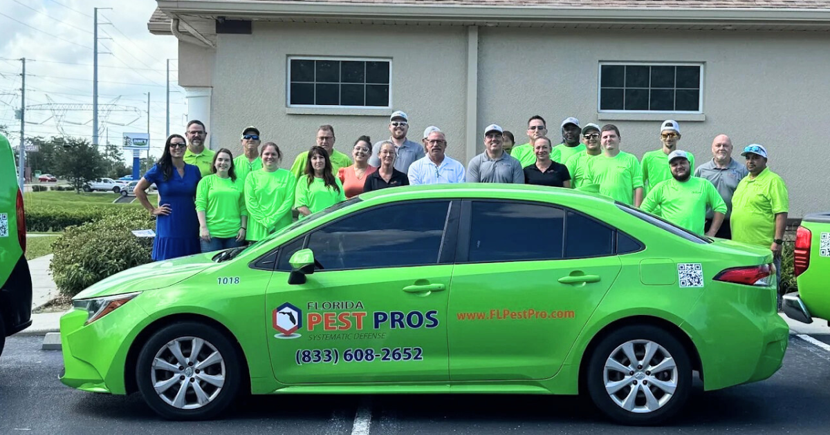Group of people standing behind a bright green car with Florida Pest Pros logo, phone number, and website on its side, outside a beige building.