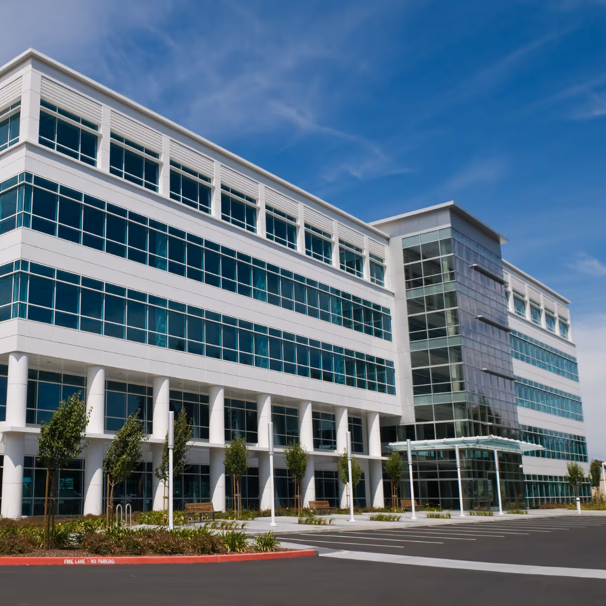 Modern commercial office building with large glass windows, white pillars, and landscaped greenery in front under a blue sky.