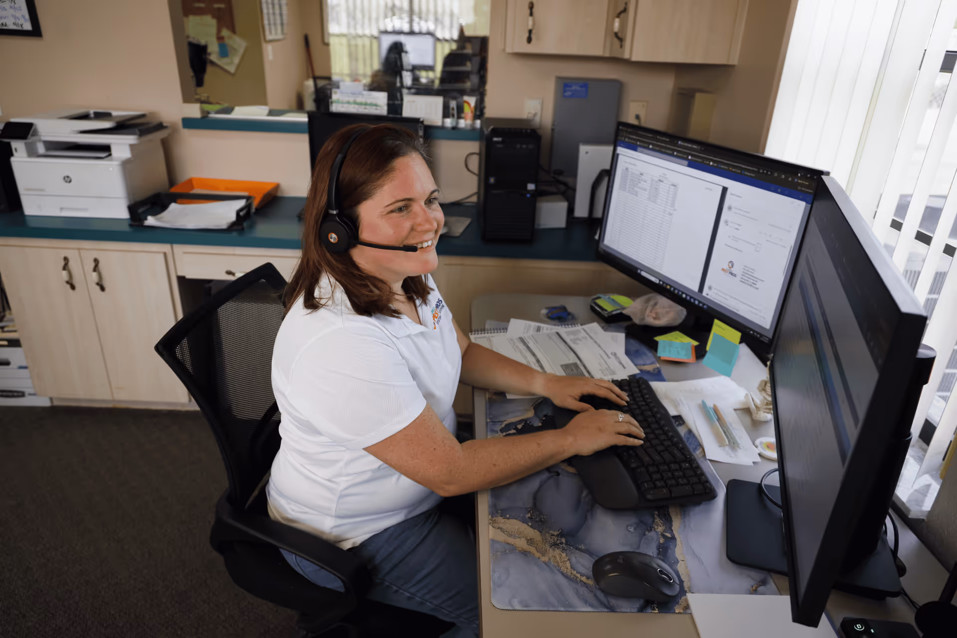 Smiling woman wearing headset working at a desk with dual monitors and documents in an office.