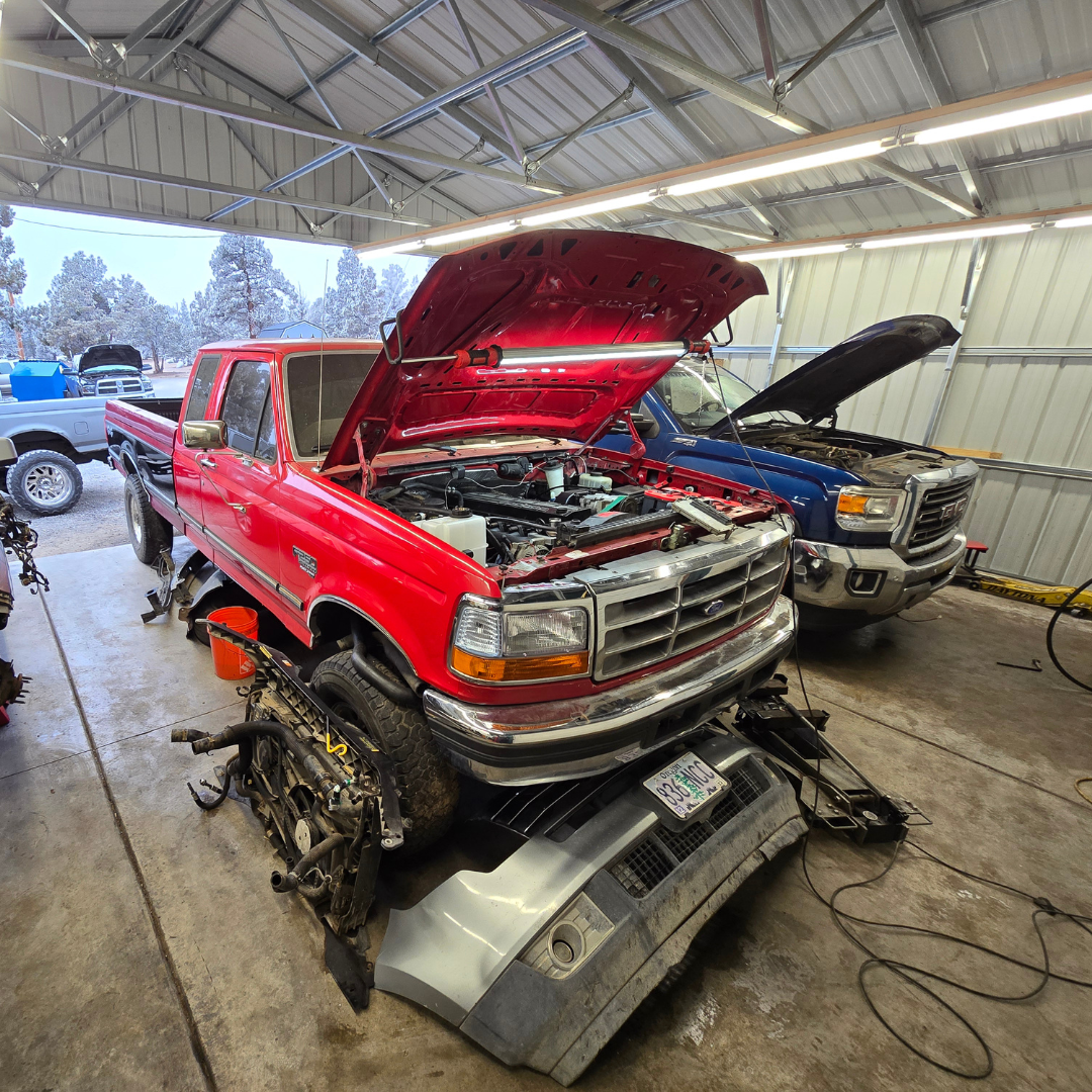 Red Ford pickup truck in a garage with its hood open and front bumper removed, next to a blue GMC truck also with its hood open.