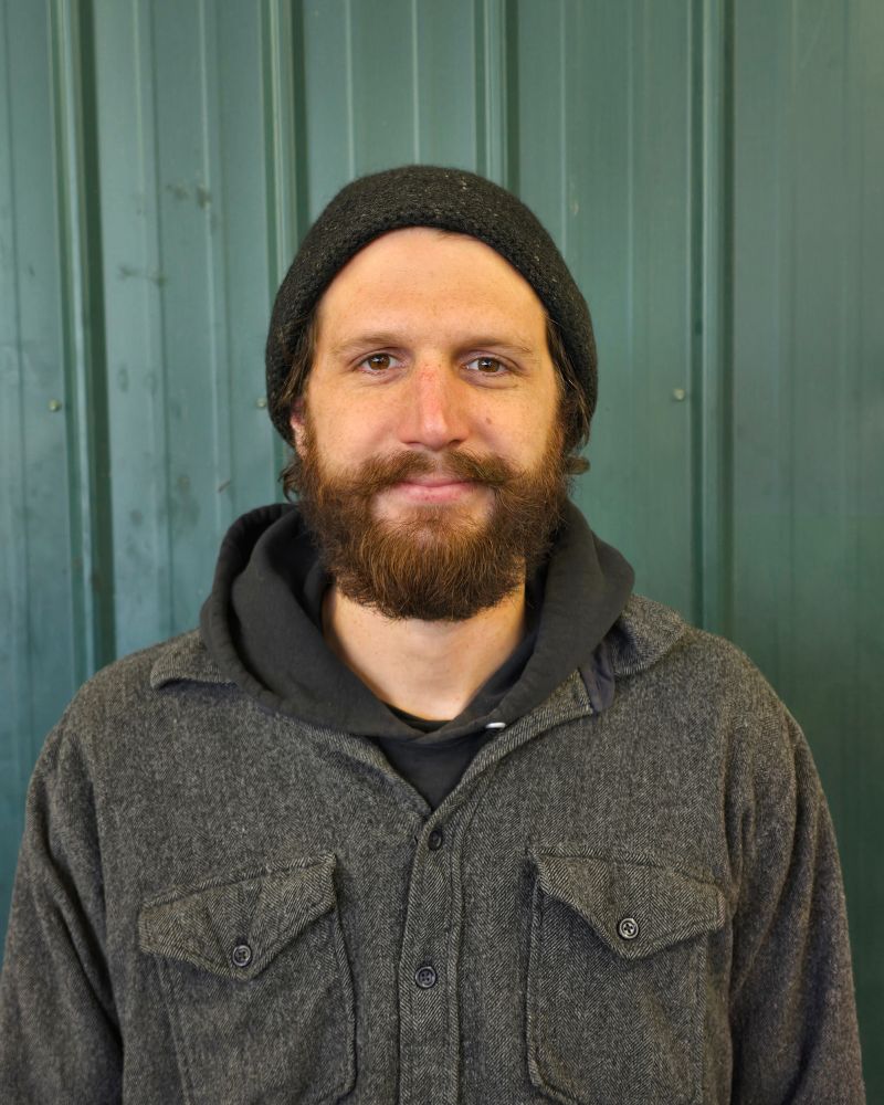 Bearded man wearing a black beanie, dark hoodie, and gray buttoned shirt standing in front of a green metal wall.