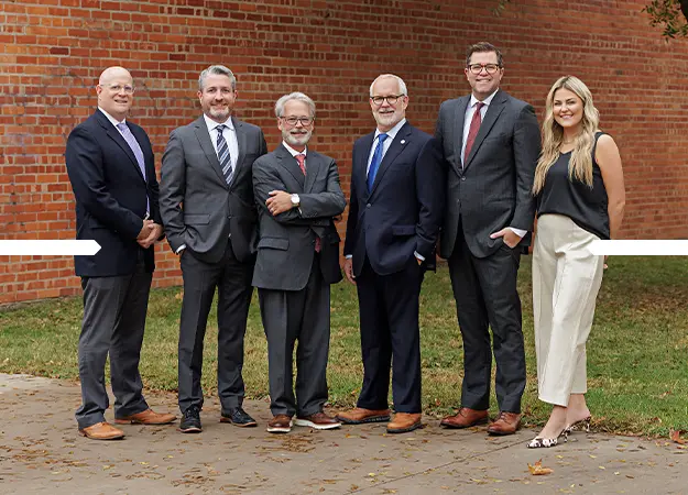 Group of six professionally dressed adults standing outdoors on a paved area with a brick wall and grass in the background.