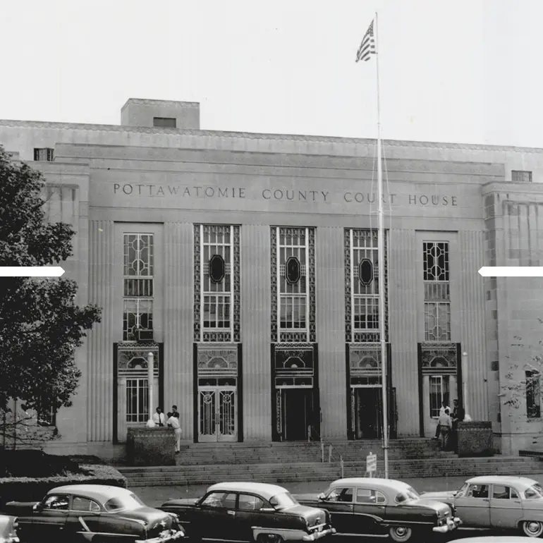Black and white photo of Pottawatomie County Court House with vintage cars parked in front and an American flag on a flagpole.